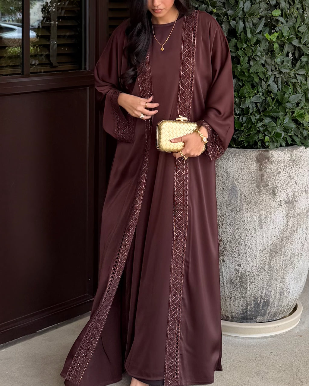 A woman wearing a long, flowing brown abaya dress with intricate lace trim, holding a small gold clutch purse, standing next to a large potted green plant.