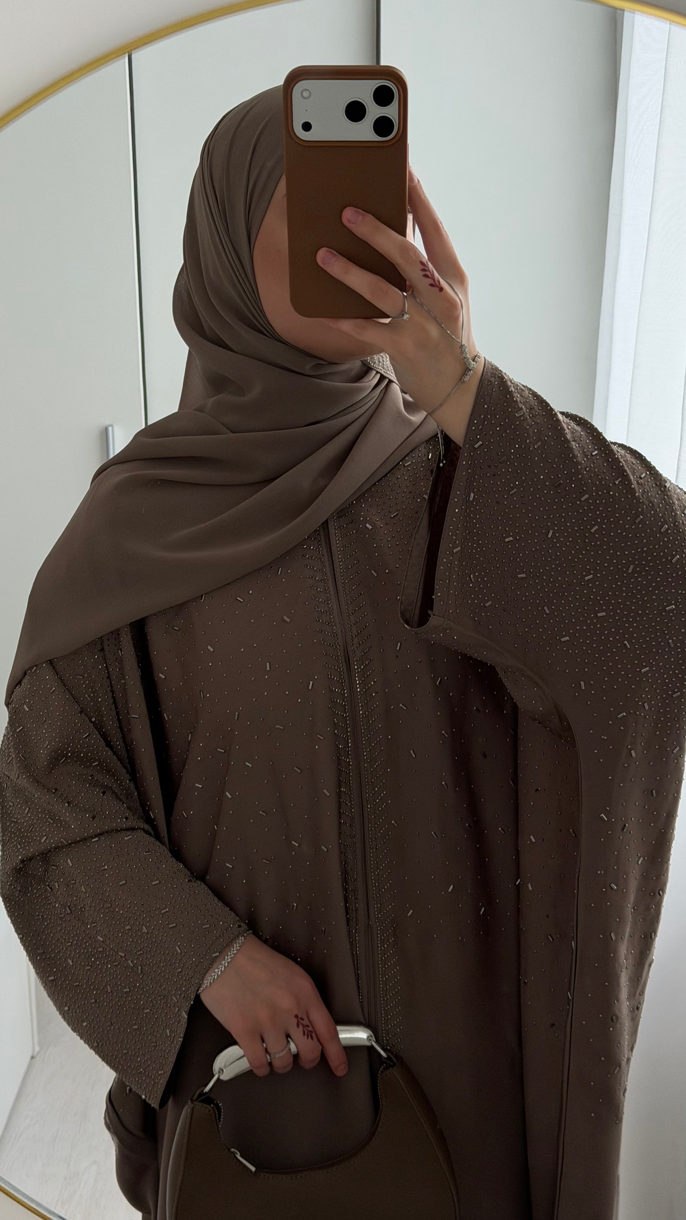 A person taking a mirror selfie wearing a brown hijab and a matching brown textured abaya with subtle silver speckles, holding a brown handbag.