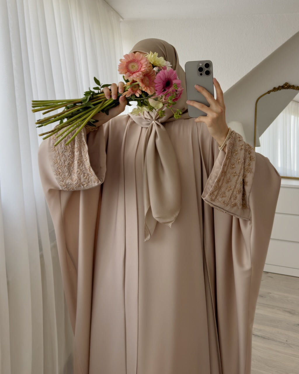 A woman takes a mirror selfie wearing a flowing beige abaya with lace-trimmed sleeves and a matching hijab. She holds a bouquet of pink, peach, and white flowers in one hand and uses a gray smartphone in the other.