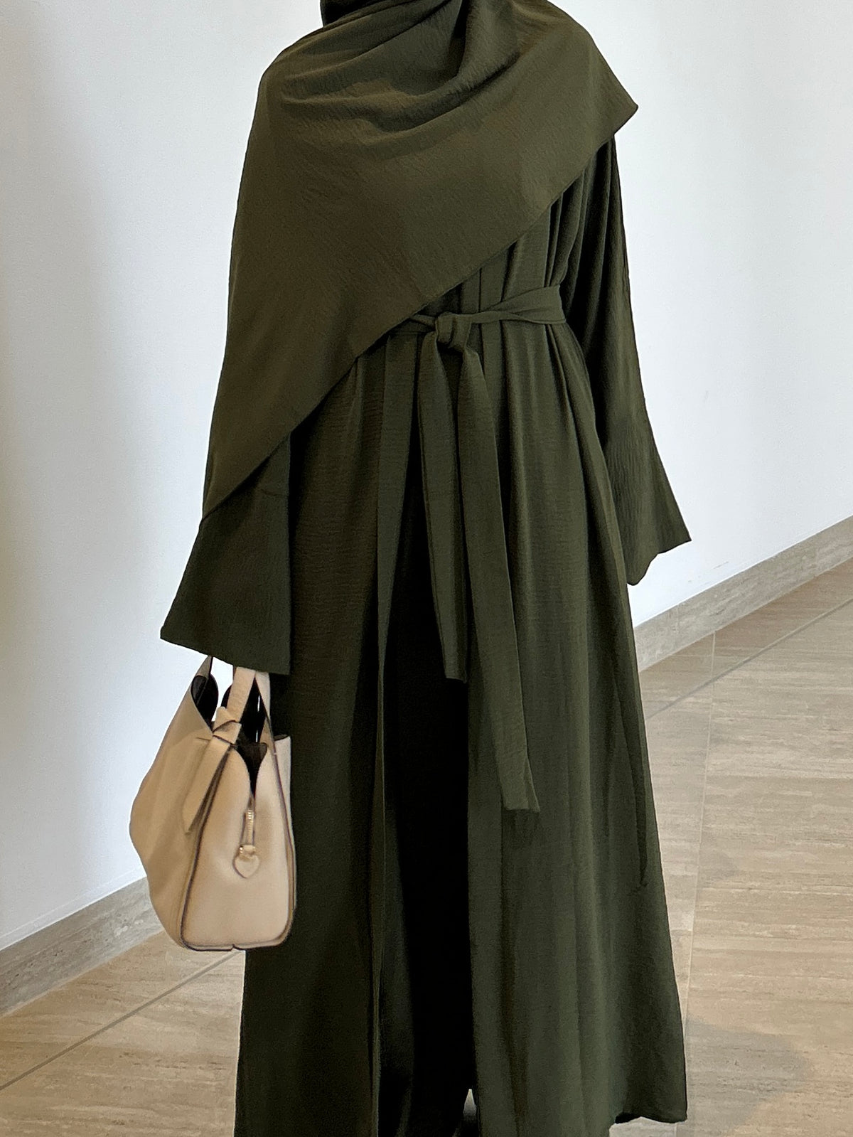 Green abaya with a beige handbag on a wooden floor against a white wall