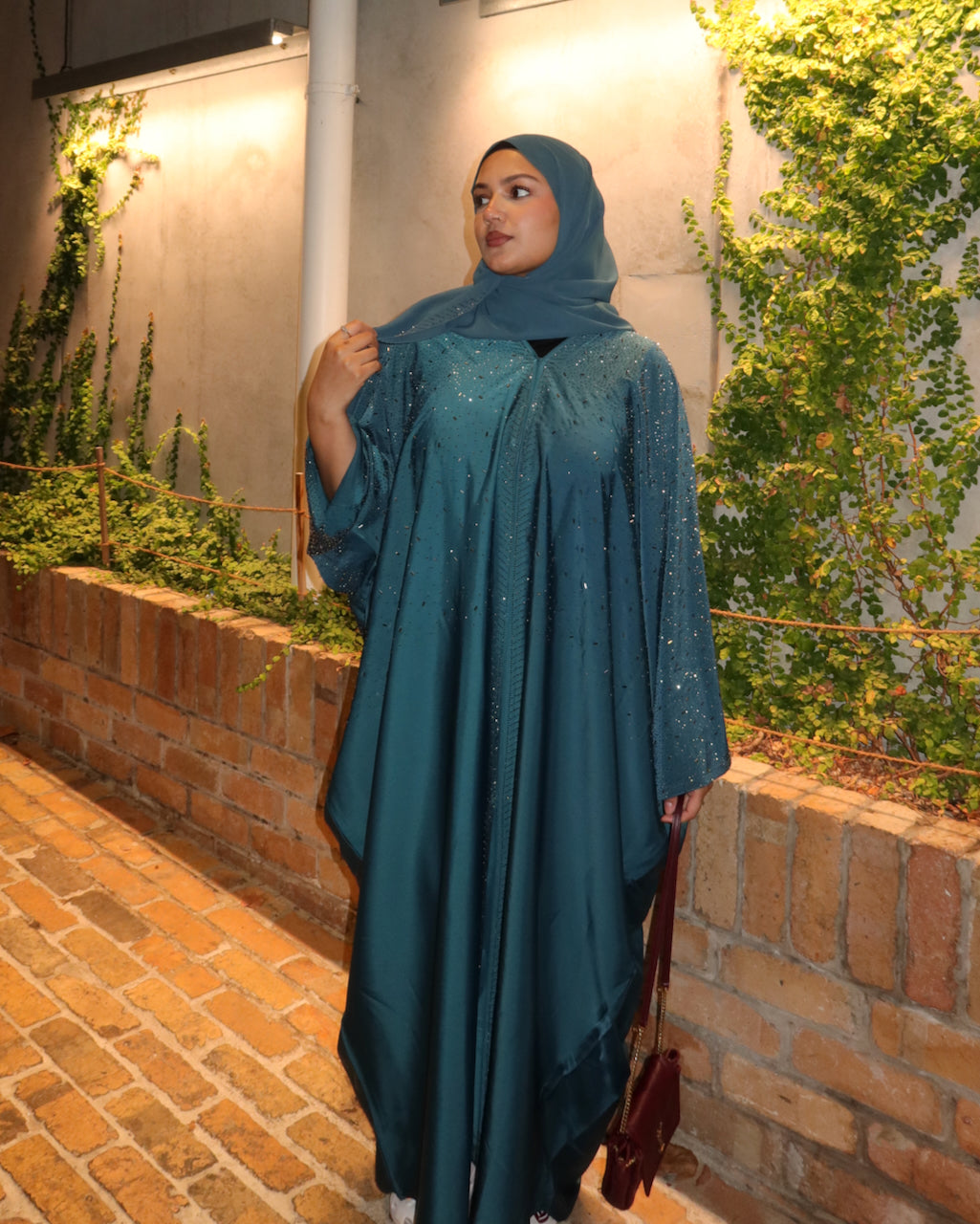 A woman wearing a teal blue-green satin abaya with subtle sequin details and a matching hijab, standing outdoors against a brick wall with green ivy.