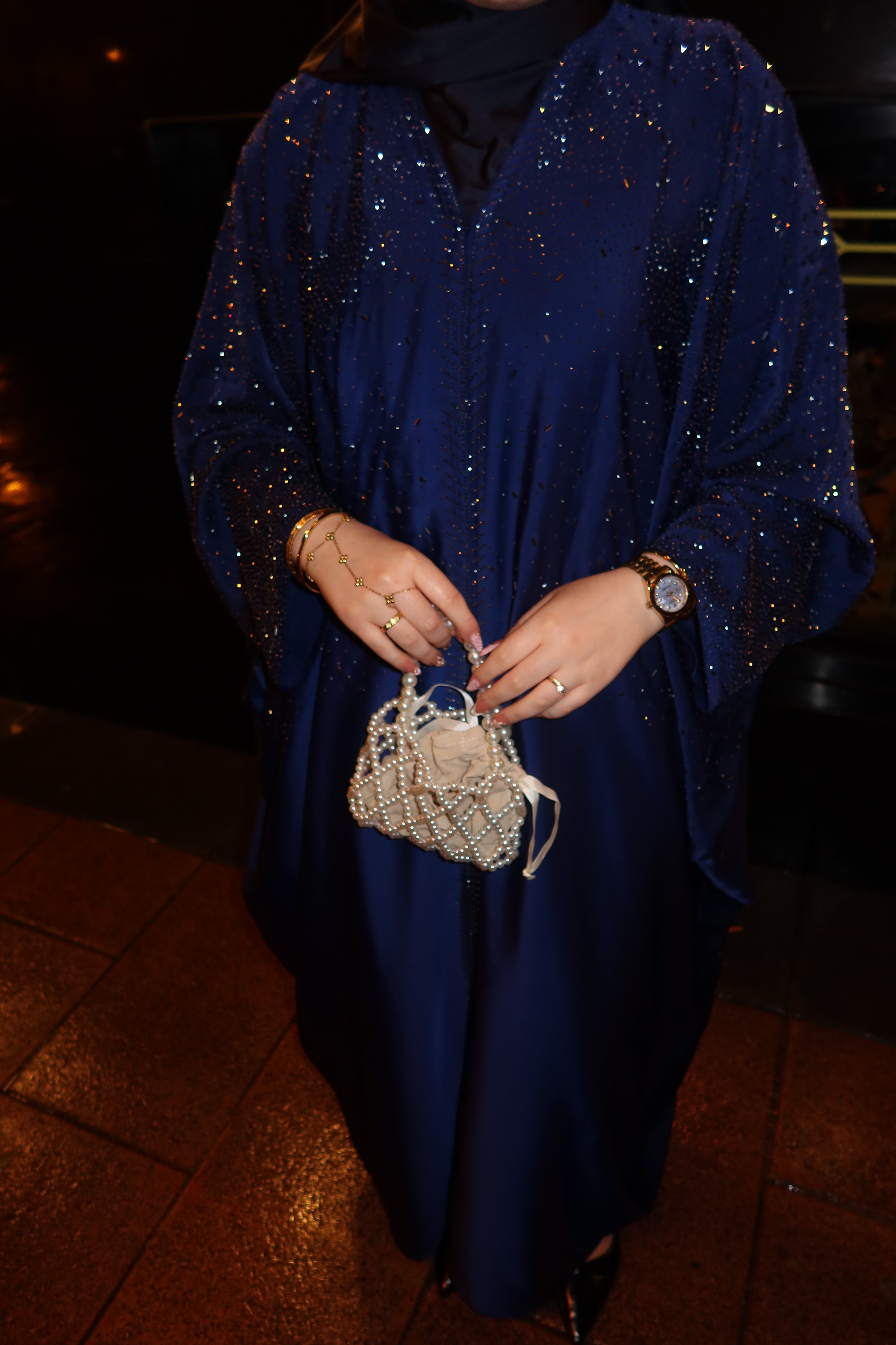 A person wearing a long, flowing navy blue abaya or dress covered in sparkling sequins, holding a small woven beige handbag. The photo is taken at night on a tiled floor, with the person's hands, wrists, and lower body visible.