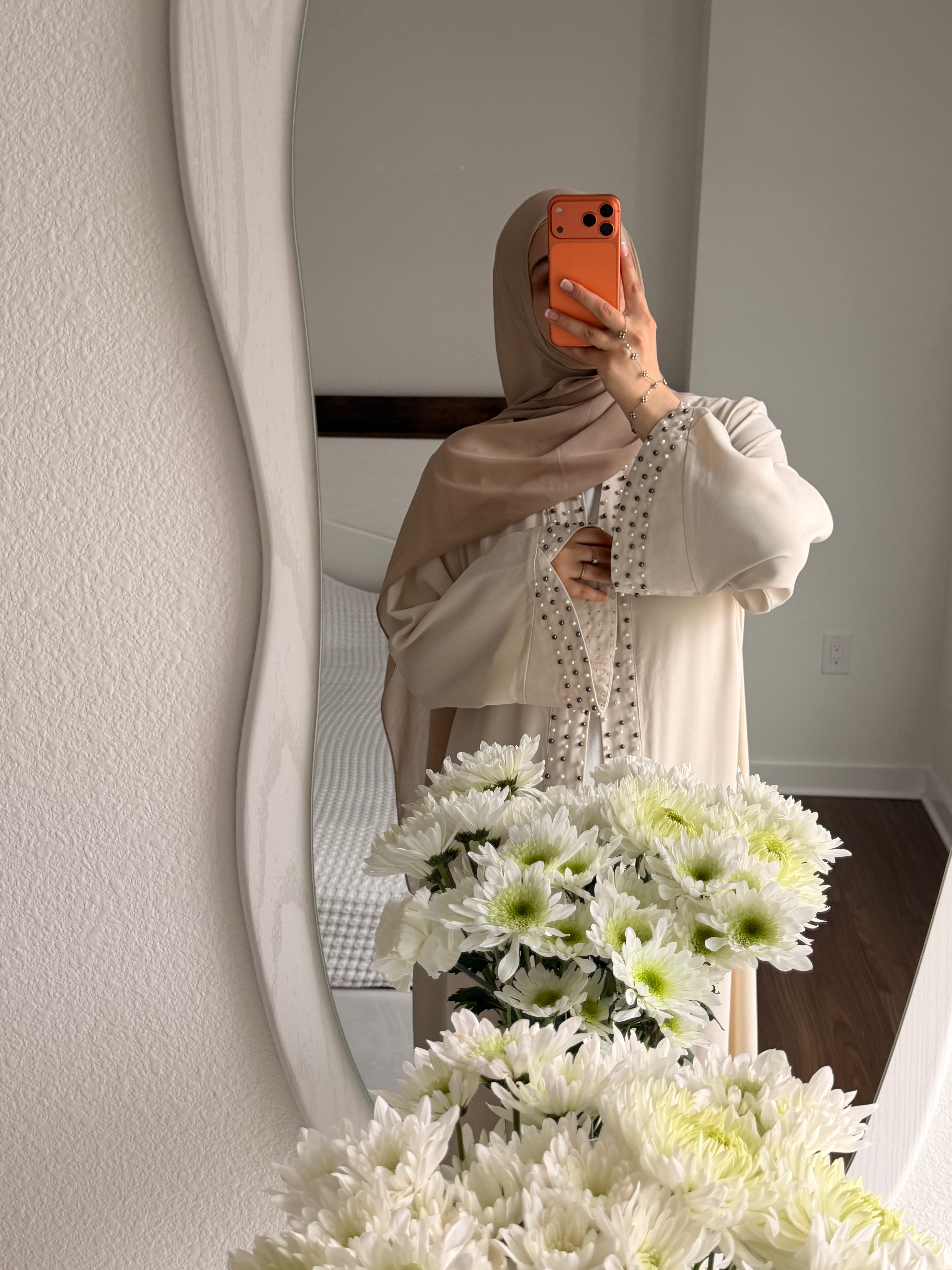 A woman wearing a beige hijab and a cream-colored long-sleeve tunic with a delicate black polka-dot patterned inner layer takes a mirror selfie in a bedroom. White flowers are in the foreground, and a bed is visible in the background.