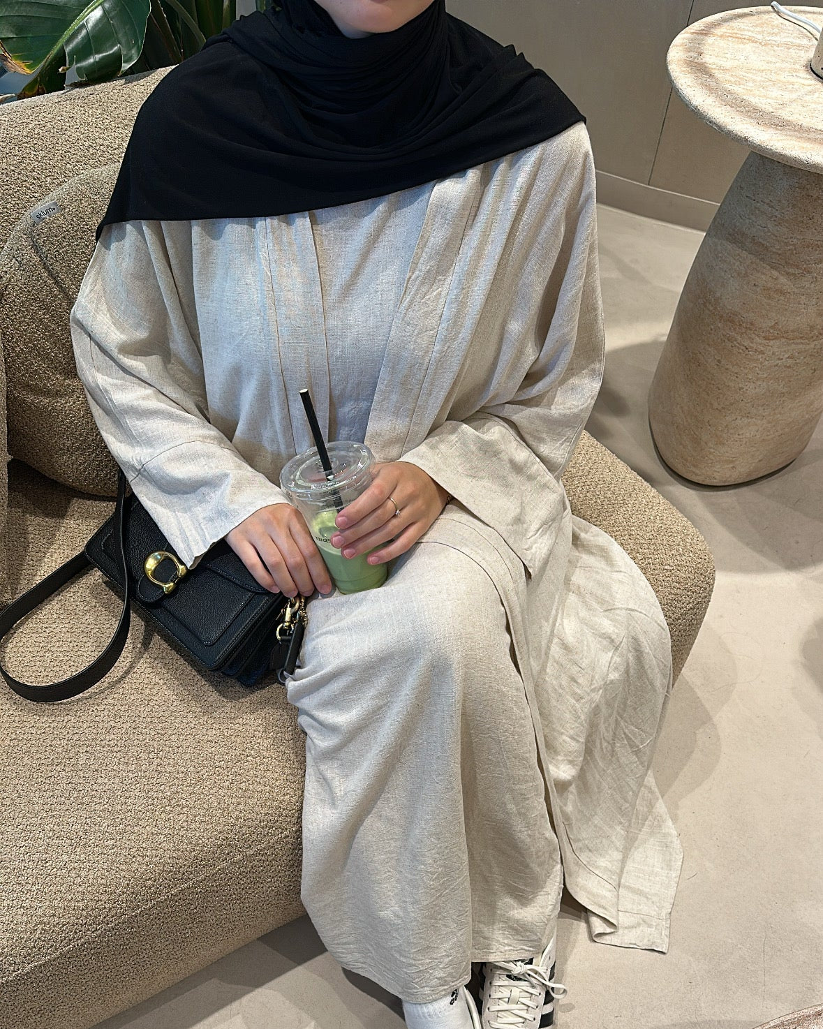 Person sitting on a couch wearing a black hijab, white outfit, and white sneakers.