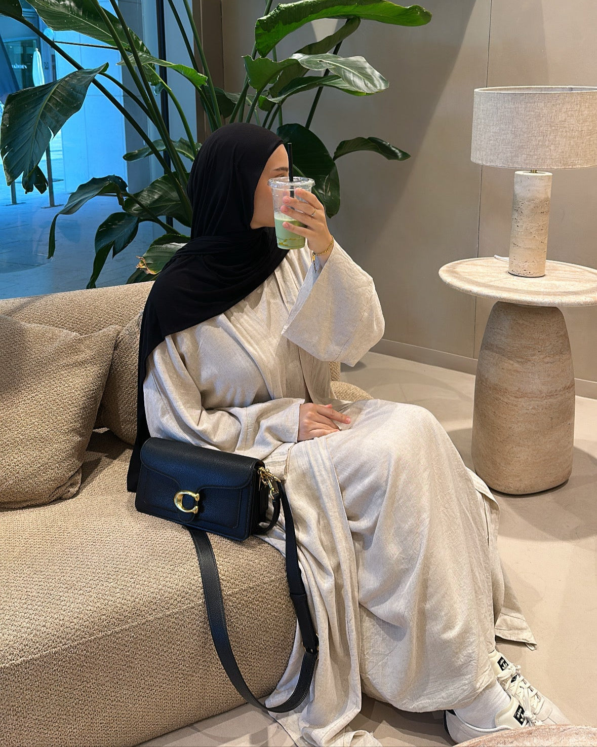 Woman in a hijab sitting on a couch with a plant and lamp in the background
