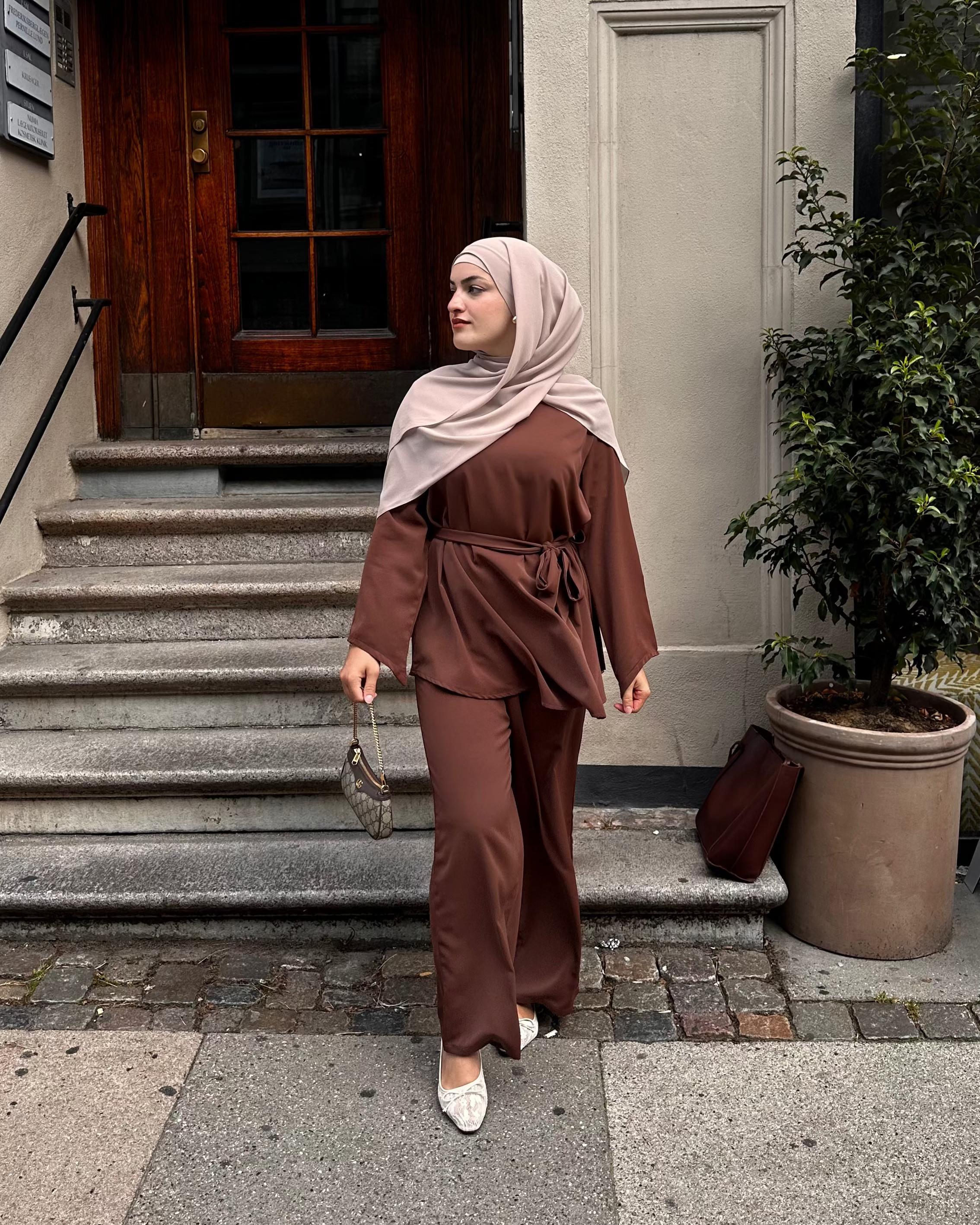 Woman in a brown outfit and hijab standing outside a building.