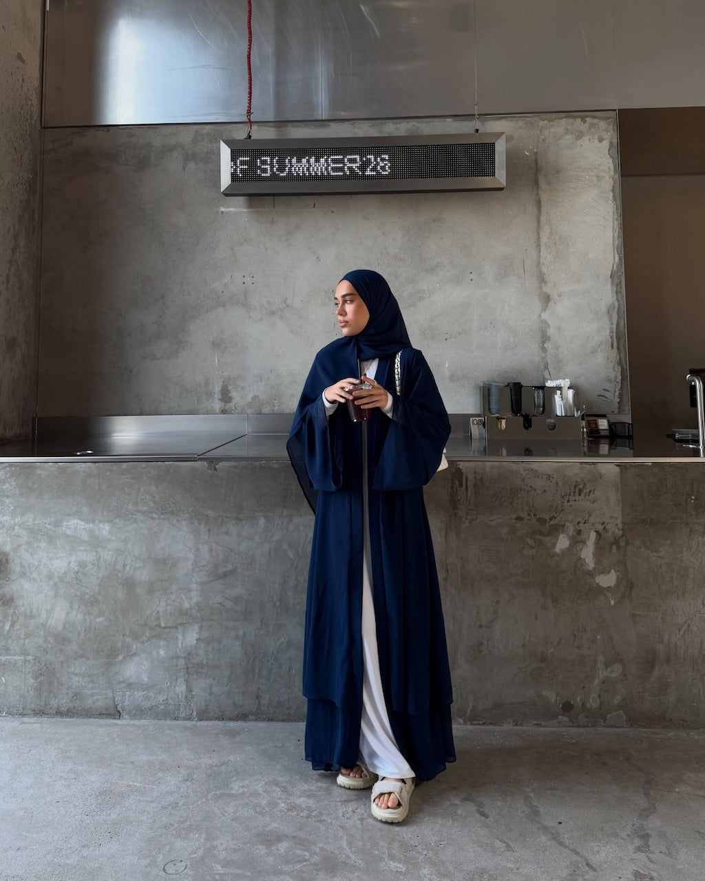 A woman wearing a long navy blue abaya with wide sleeves and a matching hijab stands in a modern industrial-style room with concrete walls and a metal counter. A sign above reads 'F SUMMER 28'.
