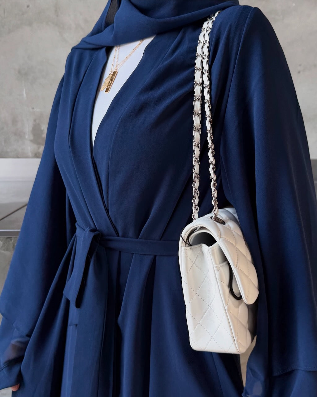 A close-up of a person wearing a navy blue draped wrap dress or abaya over a white top, styled with a white quilted shoulder bag featuring a silver chain strap.