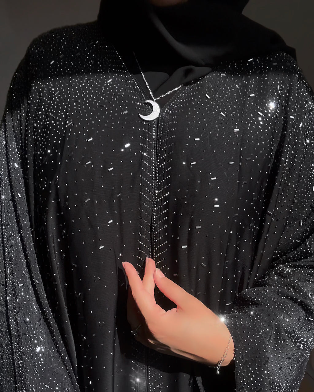 A close-up of a person wearing a black abaya or robe heavily embellished with silver rhinestones, paired with a black head covering and a silver crescent moon pendant necklace.