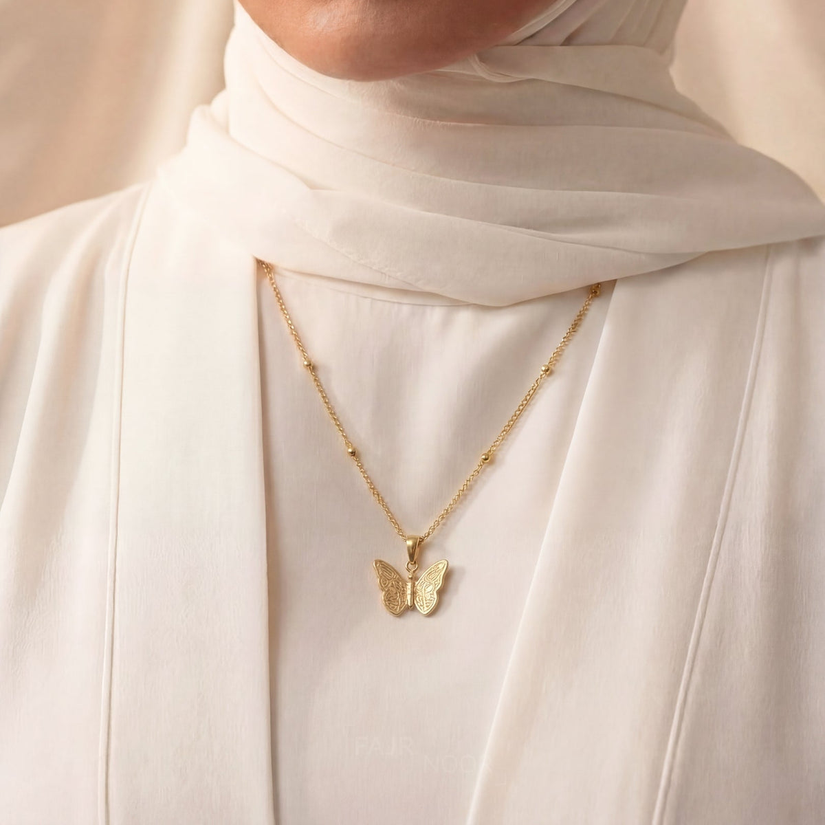 Woman wearing a gold necklace with a butterfly pendant, dressed in a white hijab and outfit.