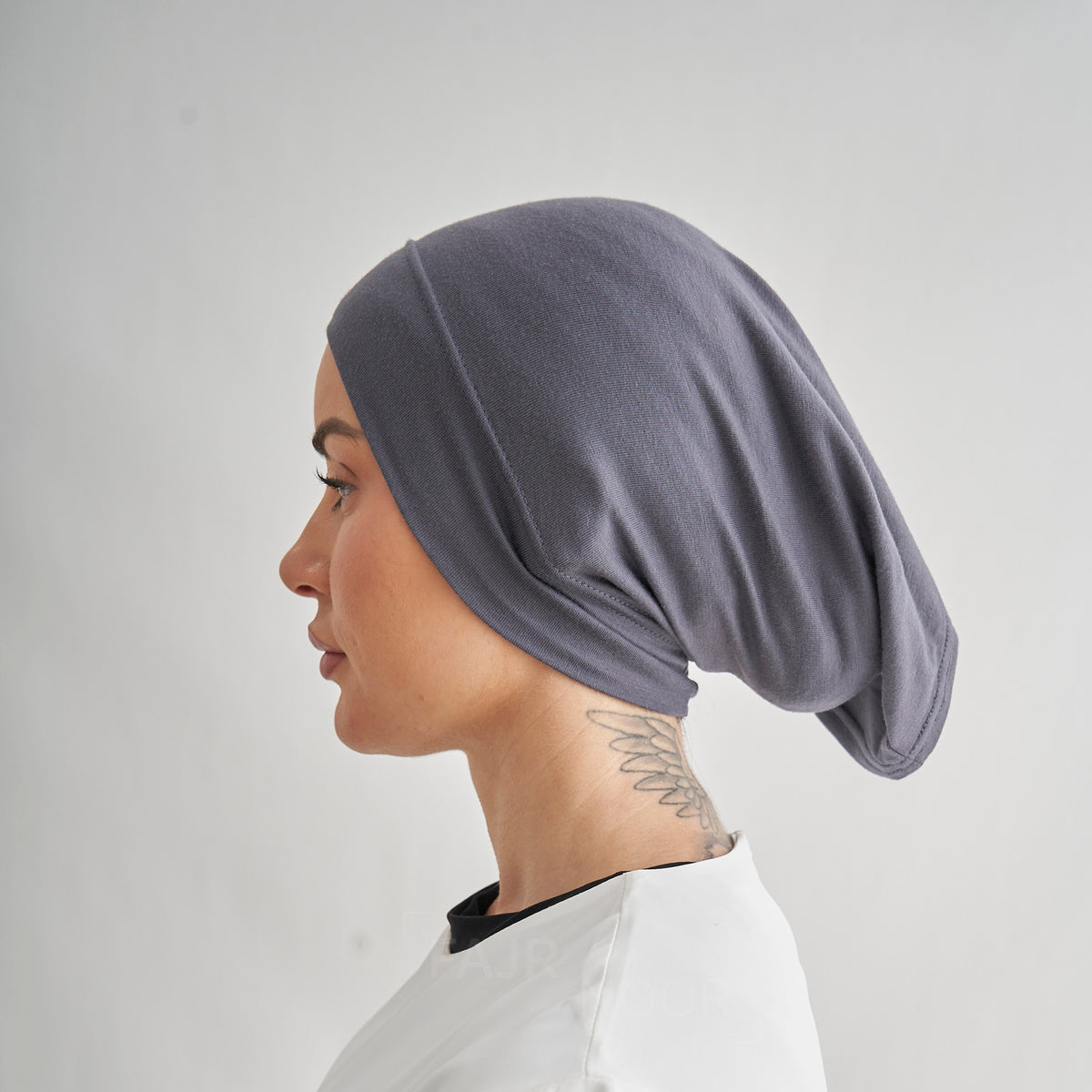 A side profile of a woman wearing a dark gray, soft-textured hijab or head covering, set against a plain light gray background.