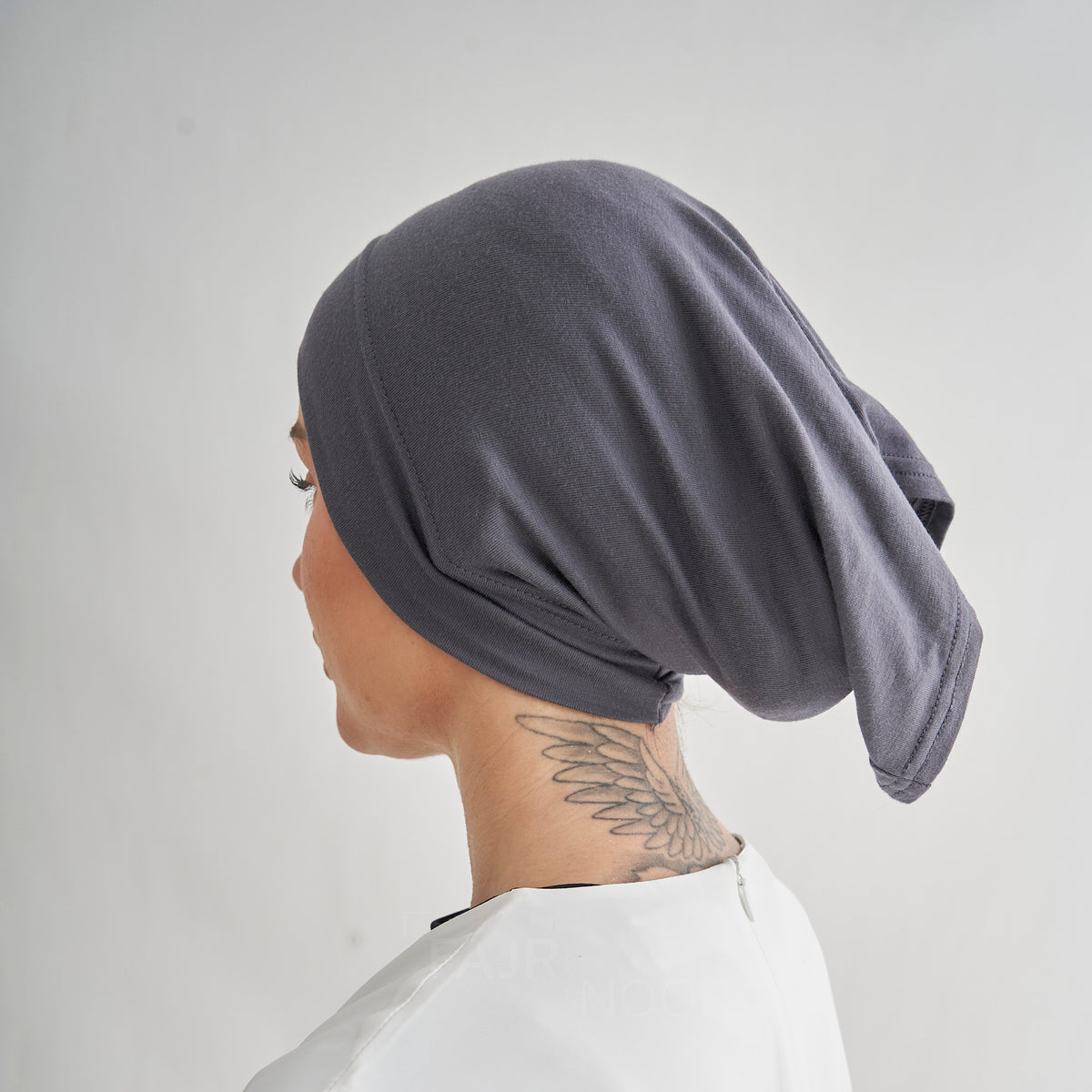 A person shown in side profile wearing a dark gray fabric head covering or hijab, photographed against a plain light gray background.