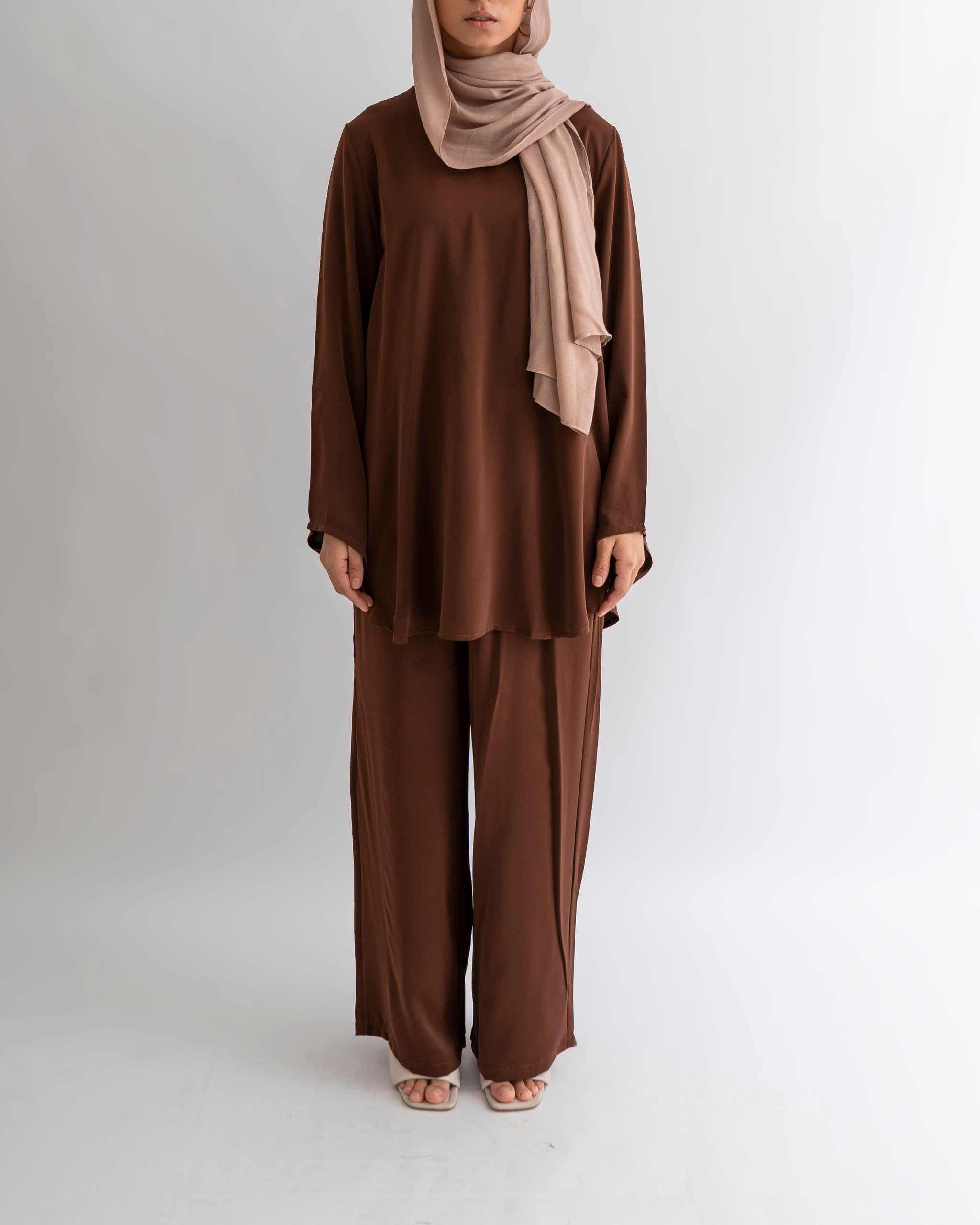 A woman wearing a brown matching set consisting of a top and trousers, with a belt, and a beige scarf.