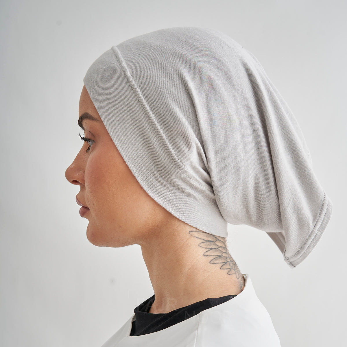 A woman shown in profile wearing a light gray knit head covering or hijab, photographed against a plain light gray background.