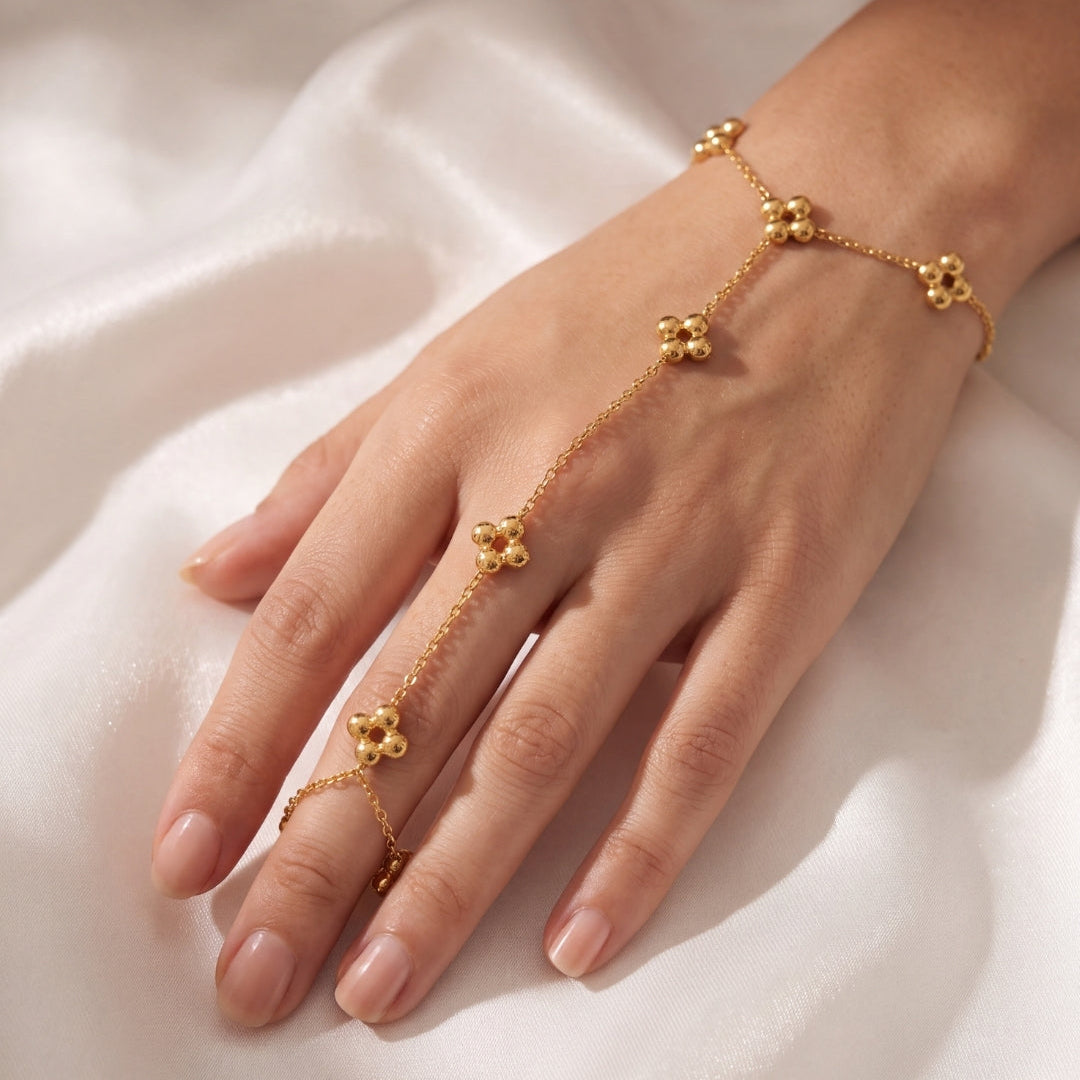 A delicate gold chain hand bracelet with small flower-like motifs worn on a person's hand, resting on soft white fabric.