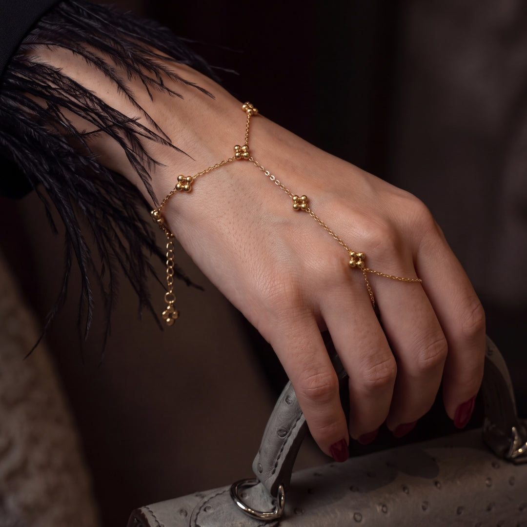 A delicate gold chain hand bracelet with small bead clusters and dangling chains, worn on a woman's hand as she holds a gray textured handbag.