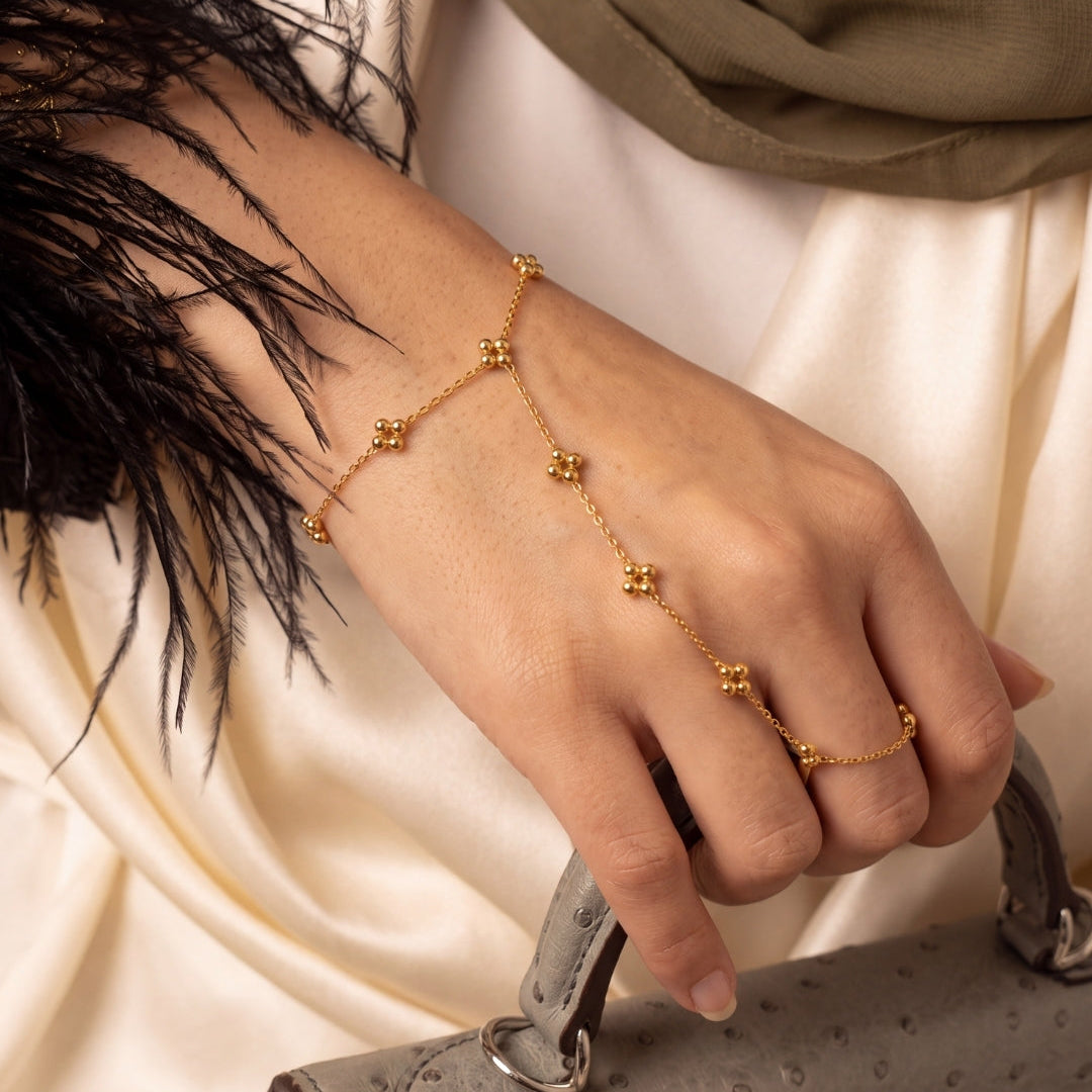 A delicate gold chain hand bracelet with small floral motifs worn on a woman's wrist, shown while she holds a gray textured handbag. She is wearing a cream satin dress with black feather embellishments and a taupe headscarf.