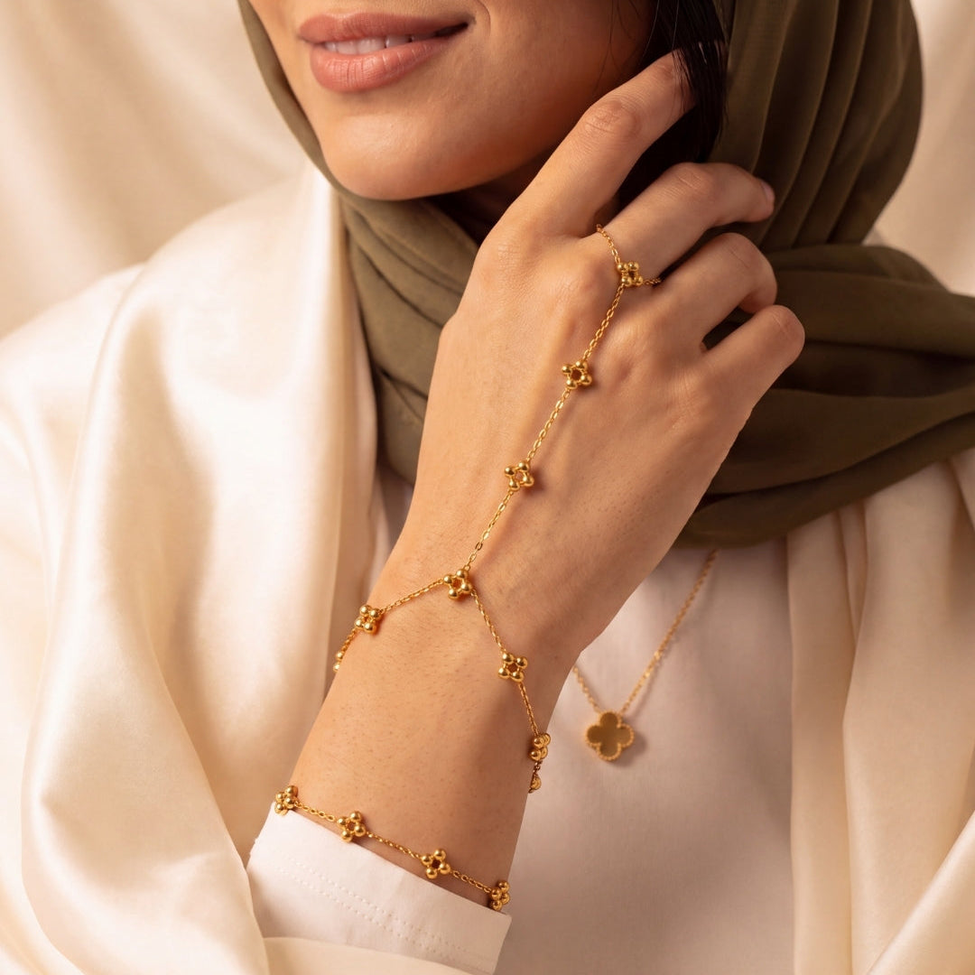 A close-up lifestyle shot of a woman wearing a delicate gold chain jewelry set, including a chain bracelet, matching ring, and necklace with clover-shaped charms, styled with a cream-colored satin robe and an olive green hijab.