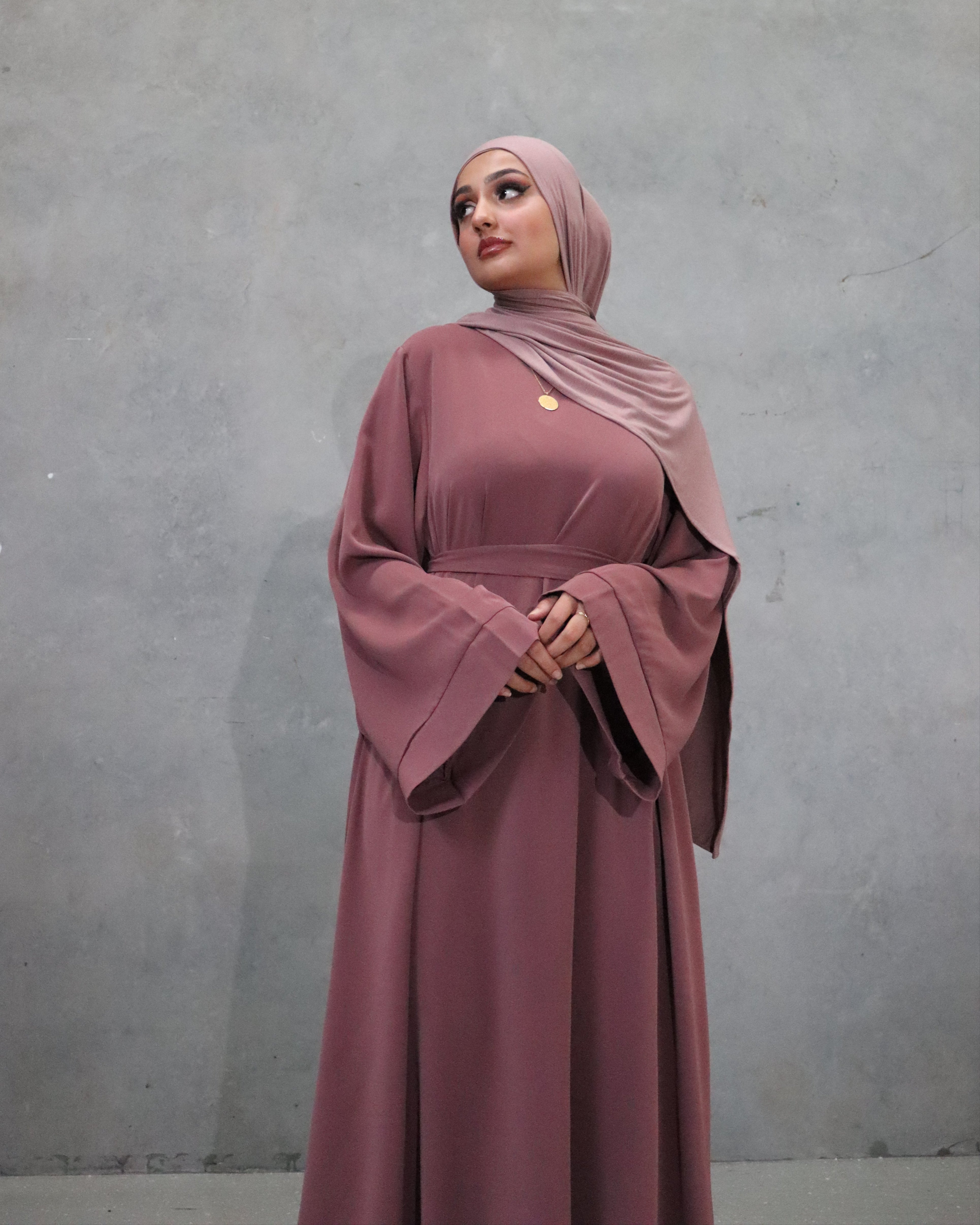 Woman wearing a mauve abaya and hijab against a gray background