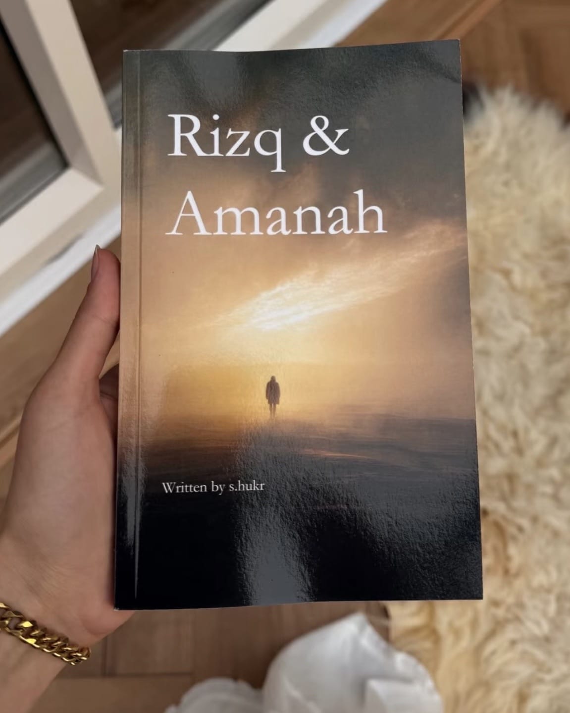 A hand holds a paperback book titled 'Rizq and Amanah' by s.hukr, featuring a cover with a sunset scene and a small silhouette of a person walking.