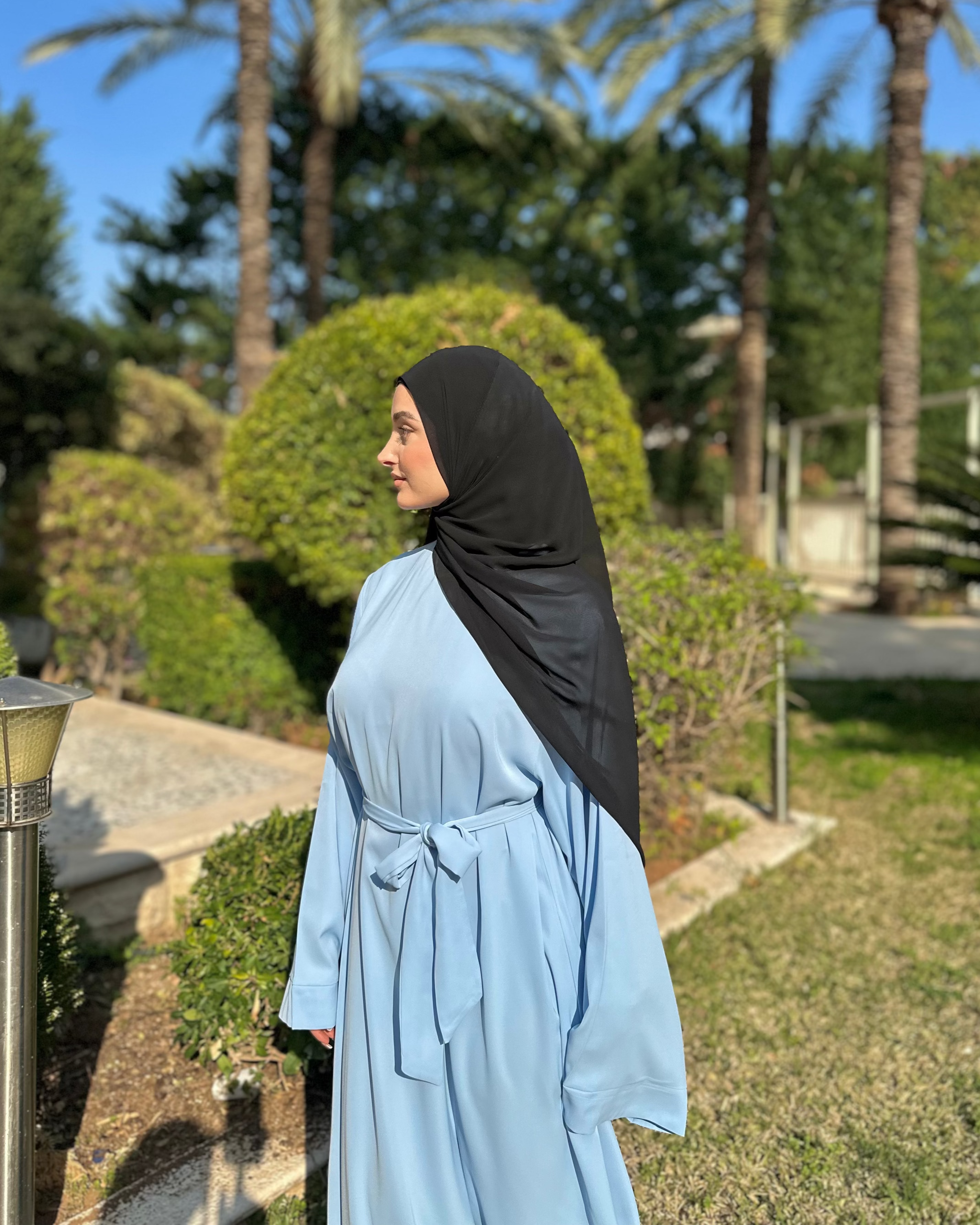 Woman in a light blue dress and black hijab standing outdoors with palm trees in the background