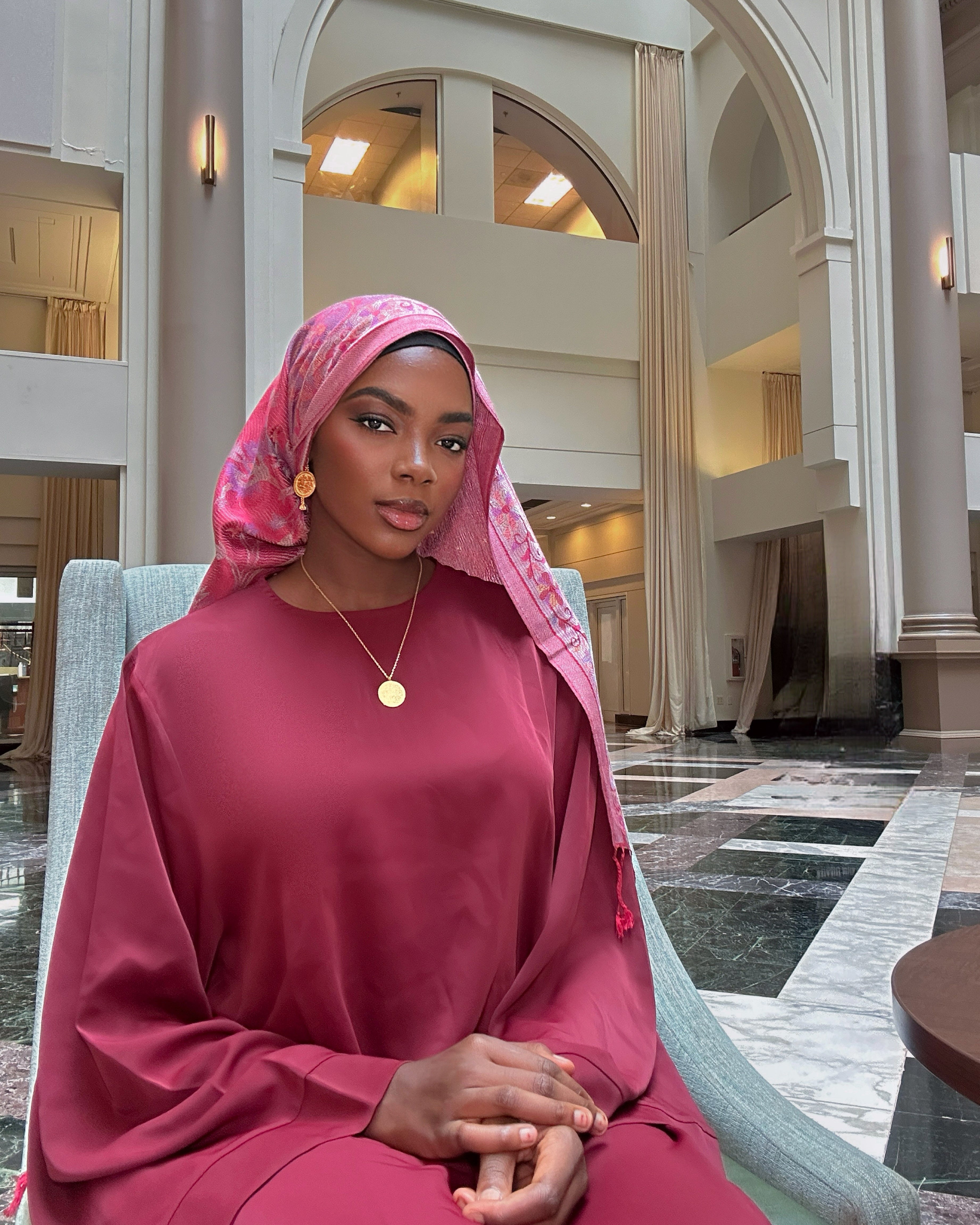 Woman in a pink outfit and headscarf sitting in a large, elegant building with high ceilings and arches.