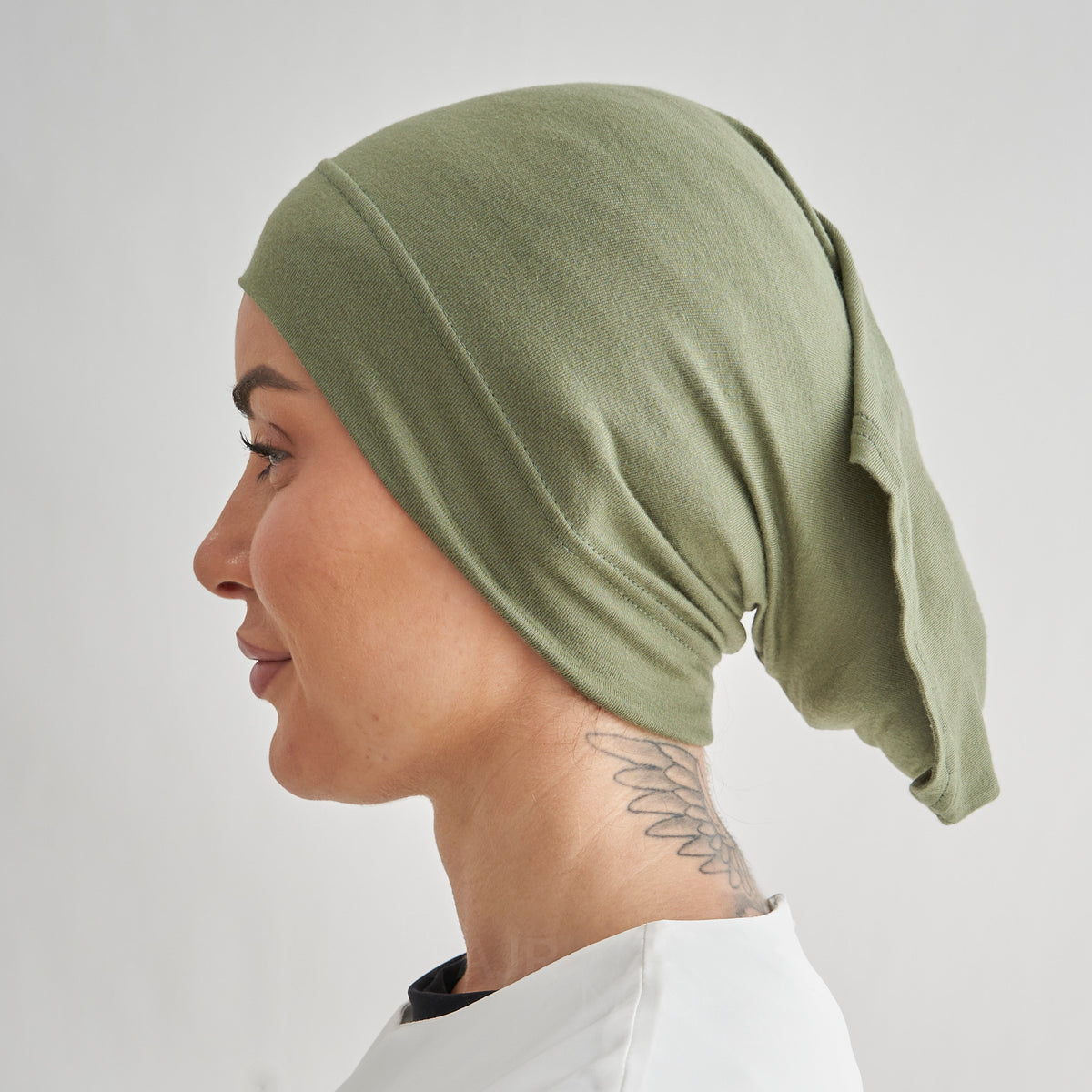 A side-profile view of a woman wearing an olive green fabric head covering or hijab against a light gray background.