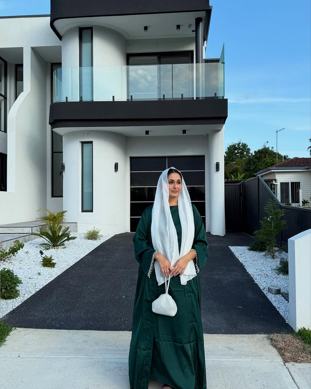 Woman in a green dress and white headscarf standing in front of a modern house.