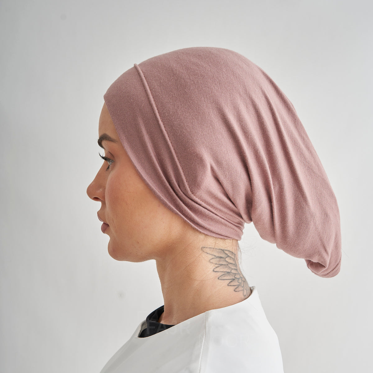 A side-profile view of a woman wearing a dusty rose pink fabric head covering or hijab, shown against a plain light gray background.