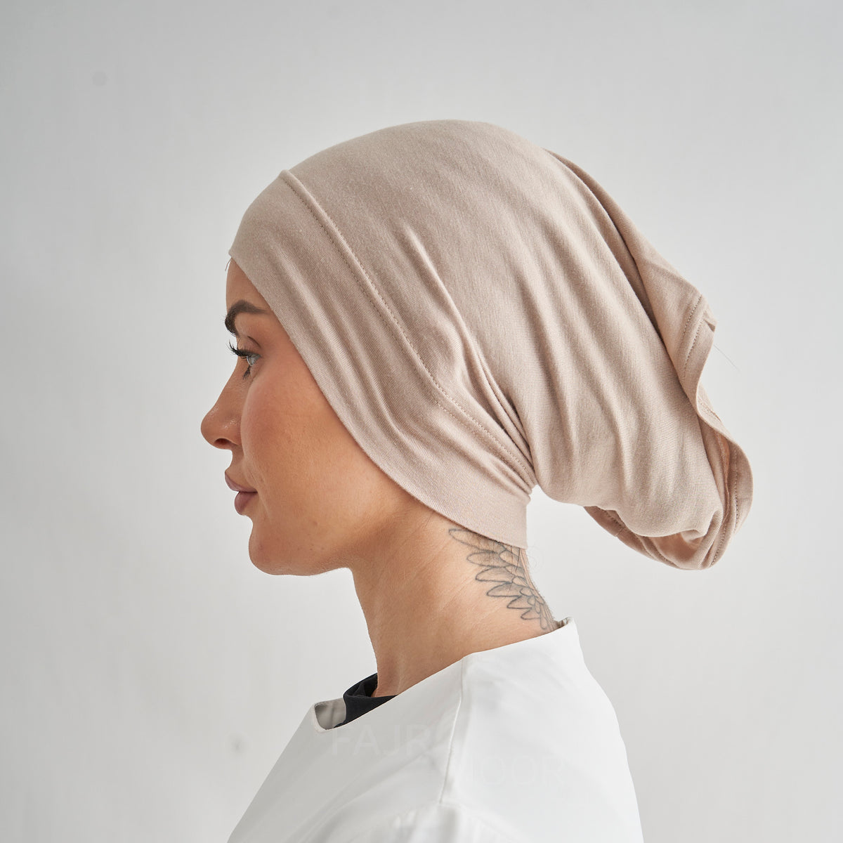 A side-profile view of a woman wearing a soft, draped beige fabric head covering or hijab against a plain light gray background.