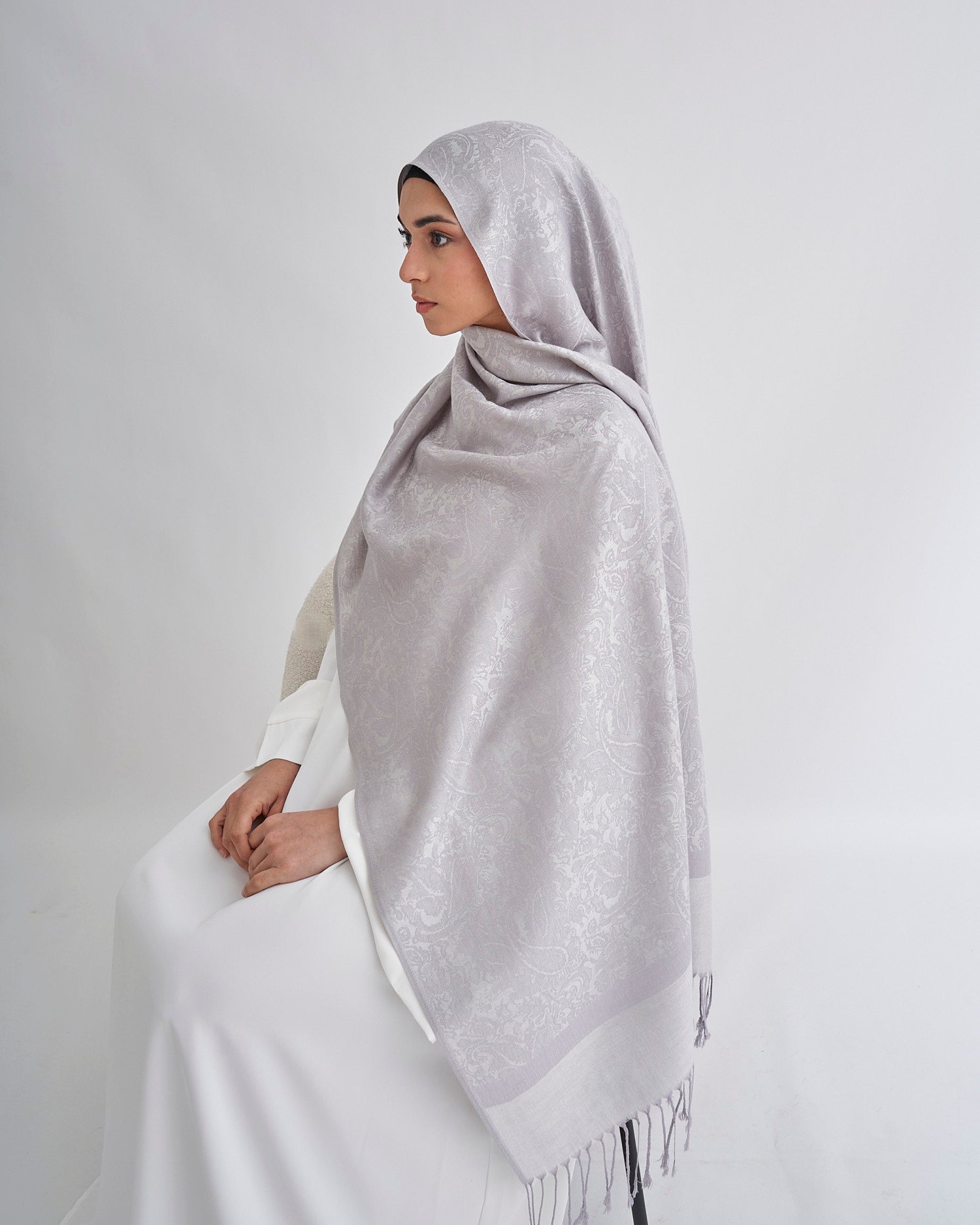 Woman wearing a light gray hijab and white dress on a plain background