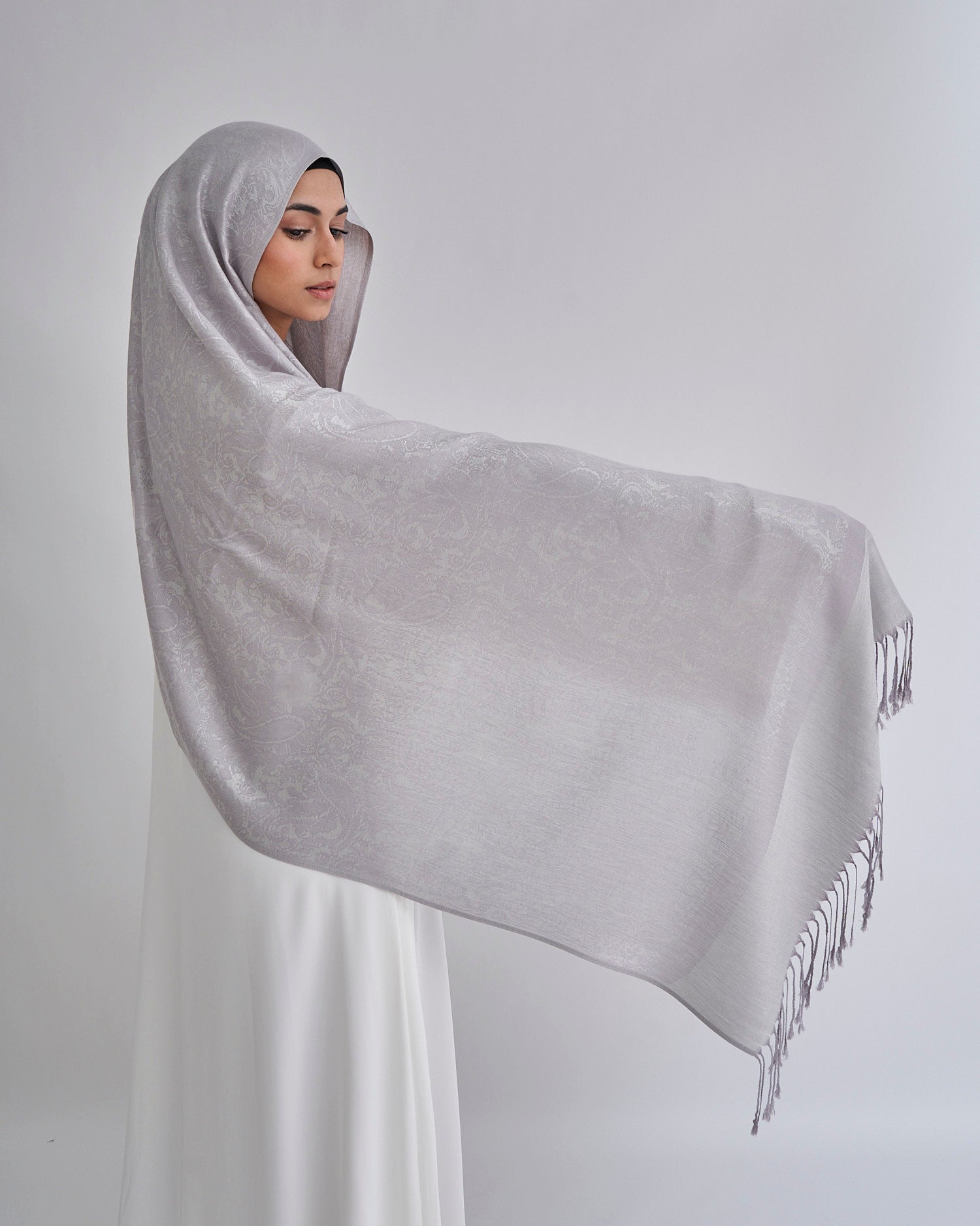 Woman draped in a large gray shawl with tassels on a plain background