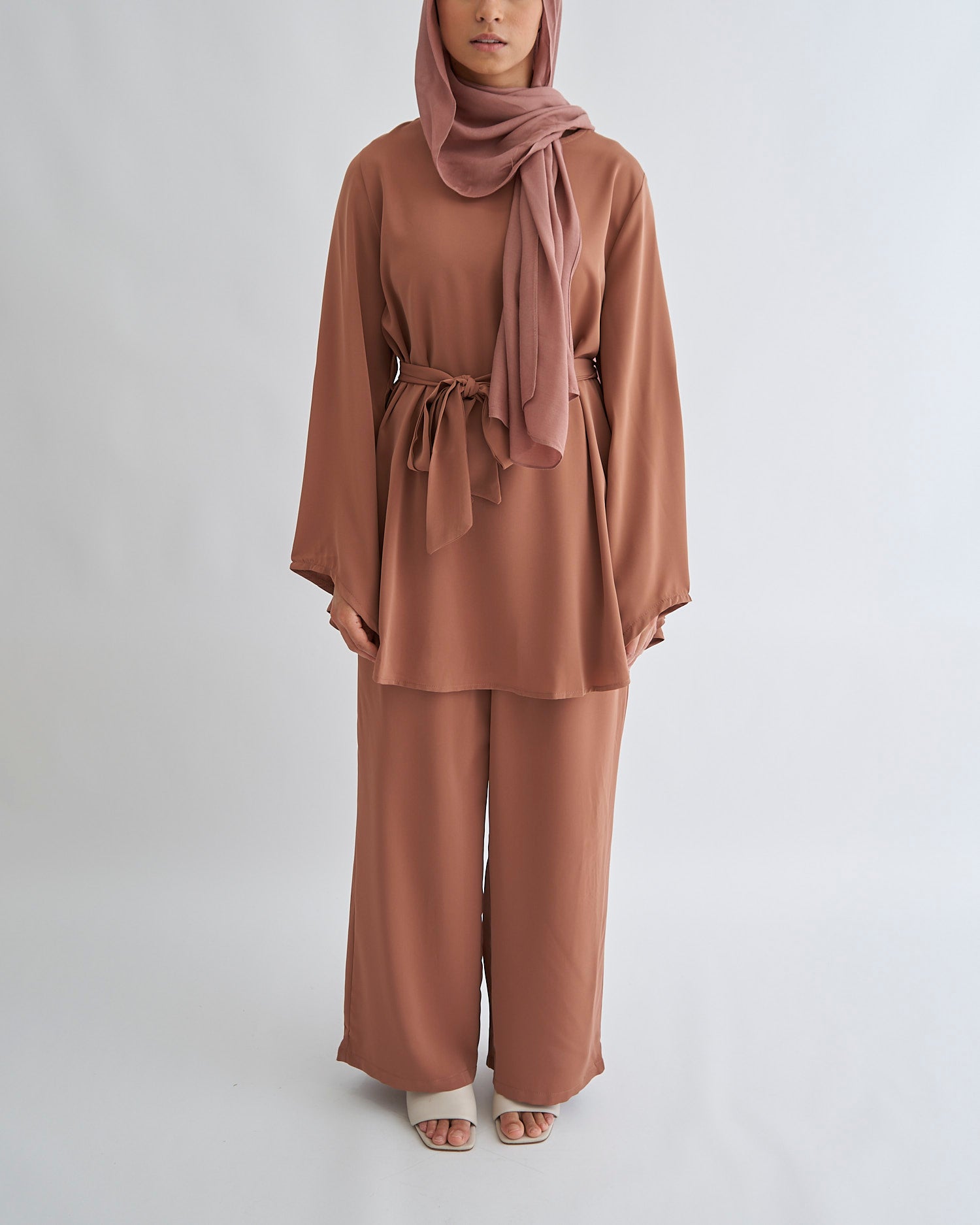 A woman wearing a brown outfit consisting of a top and wide trousers with a belt, including a matching headscarf.