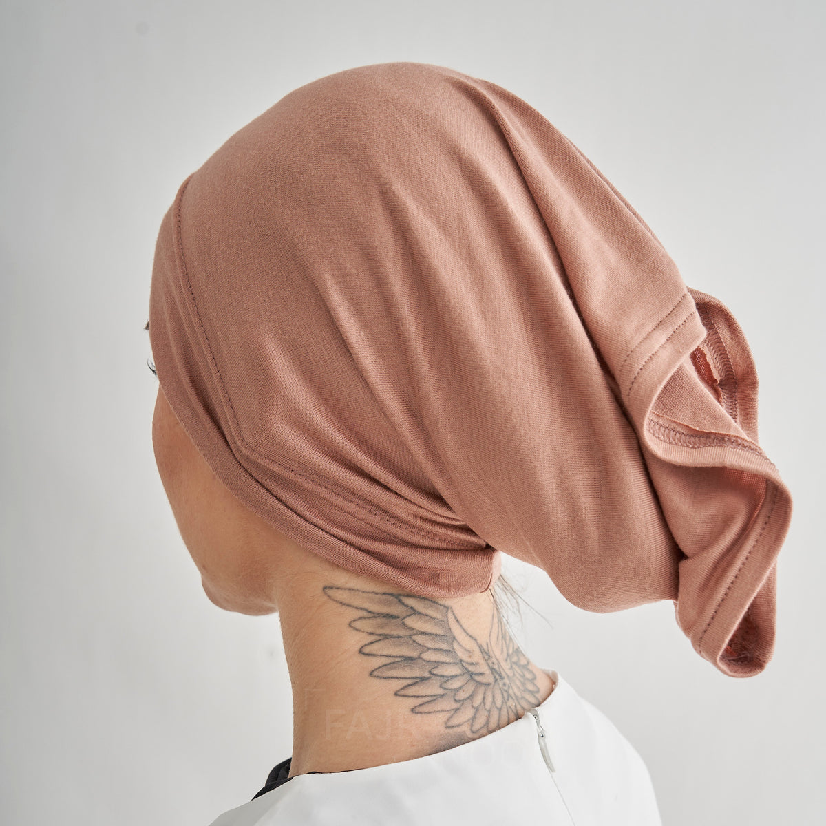 A close-up side view of a person wearing a soft, draped dusty rose head covering or hijab, with a wing tattoo visible on the neck, against a plain light gray background.