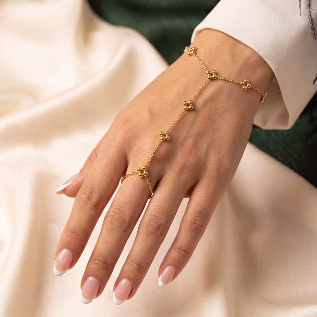 A delicate gold chain hand chain worn on a woman's hand, featuring small gold flower-like beads connected by a fine chain, paired with a matching gold ring. The hand is styled with a white long-sleeve shirt and manicured nails, set against a soft cream satin fabric and a dark green background.