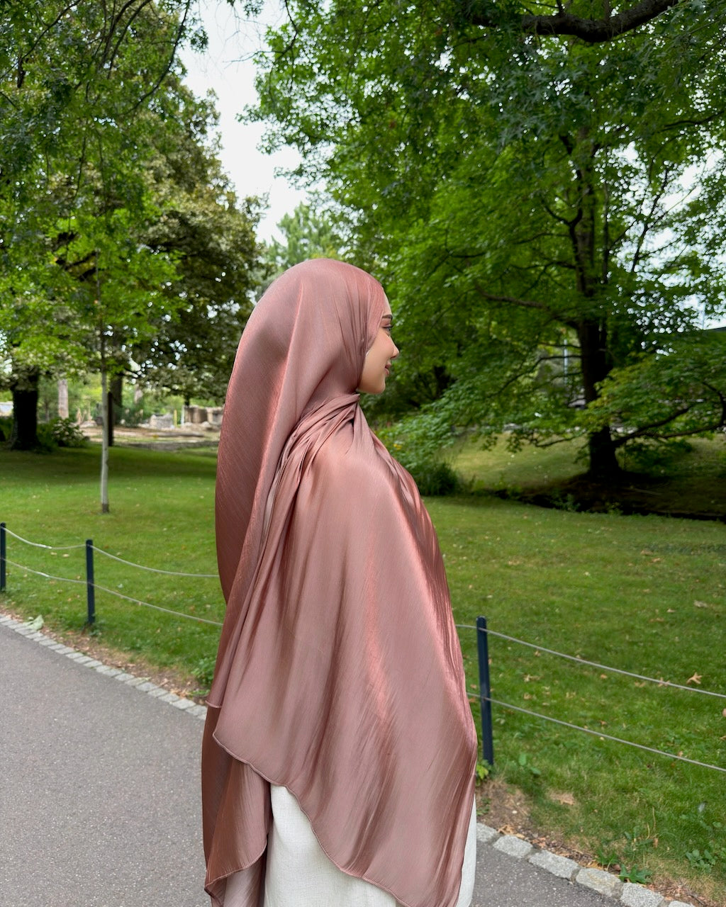 A woman wearing a dusty rose satin hijab and white pants stands in profile on a paved path in a park with green grass and trees.