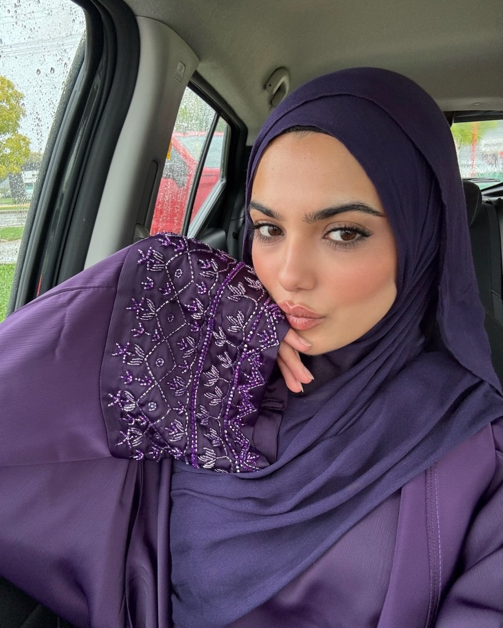 A woman wearing a deep purple hijab and matching abaya with intricate purple beaded embroidery on the sleeve, sitting inside a car.