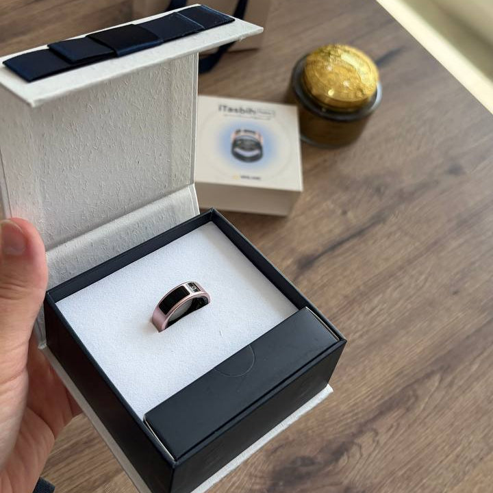 A hand holds an open black and white jewelry box containing a slim metallic ring. The scene is set on a wooden table with a shopping bag, a small box, and a gold-lidded container in the background.