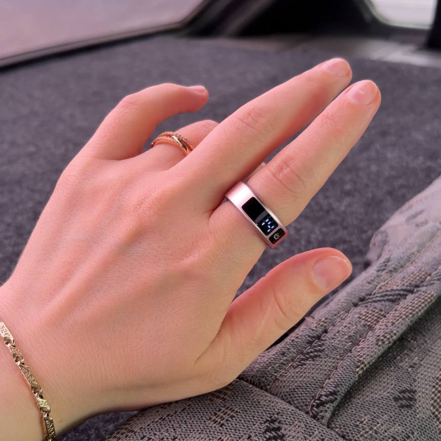 A close-up of a hand wearing a silver ring with a black rectangular inlay and the initials 'NK' on the side, set against the backdrop of a car interior.