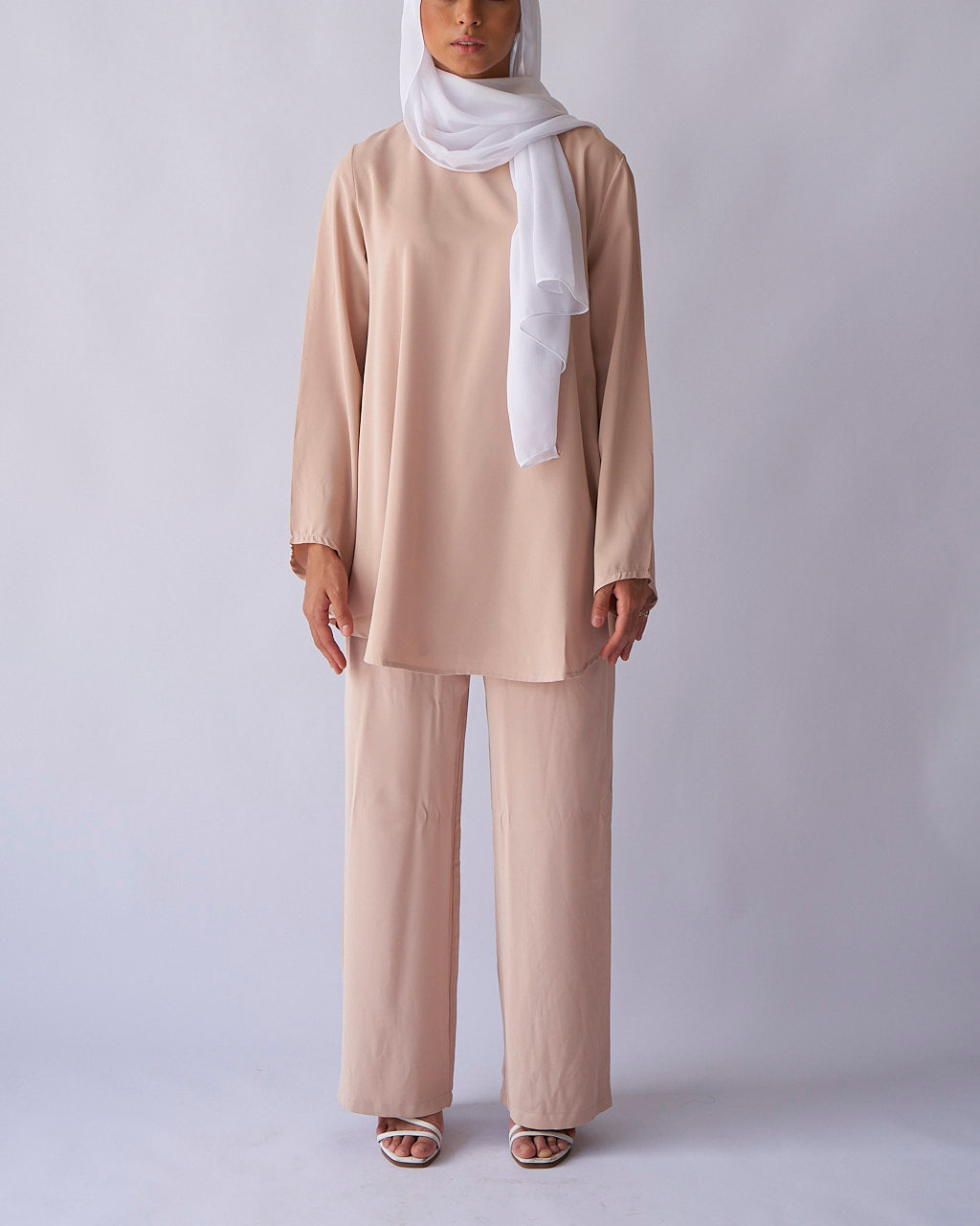 A model wearing a cream-colored top and matching wide trousers with a belt, complete with a white headscarf.