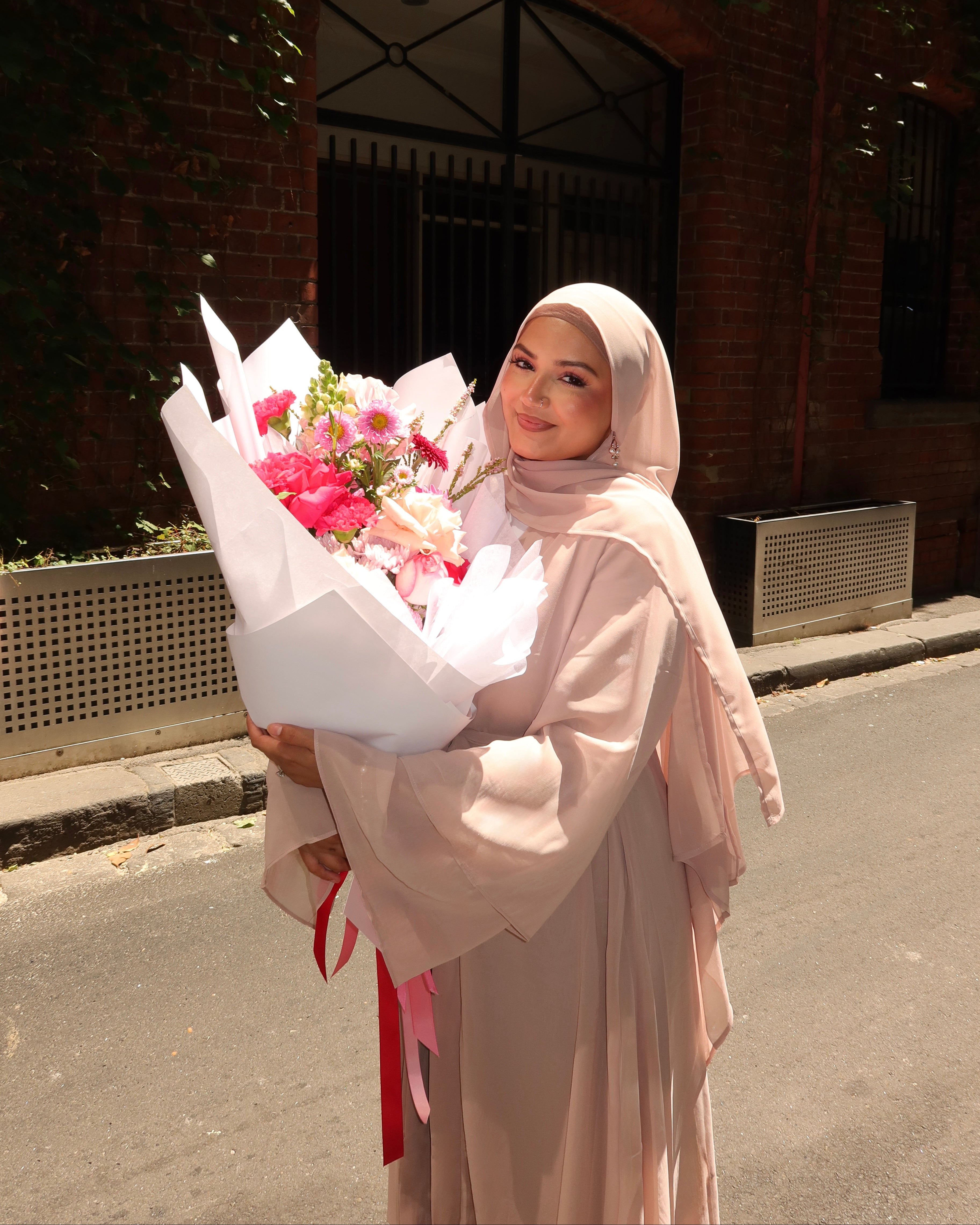 Hijabi Woman wearing Nude Aisha Chiffon Abaya holding a bouquet of flowers on a street with a brick building in the background