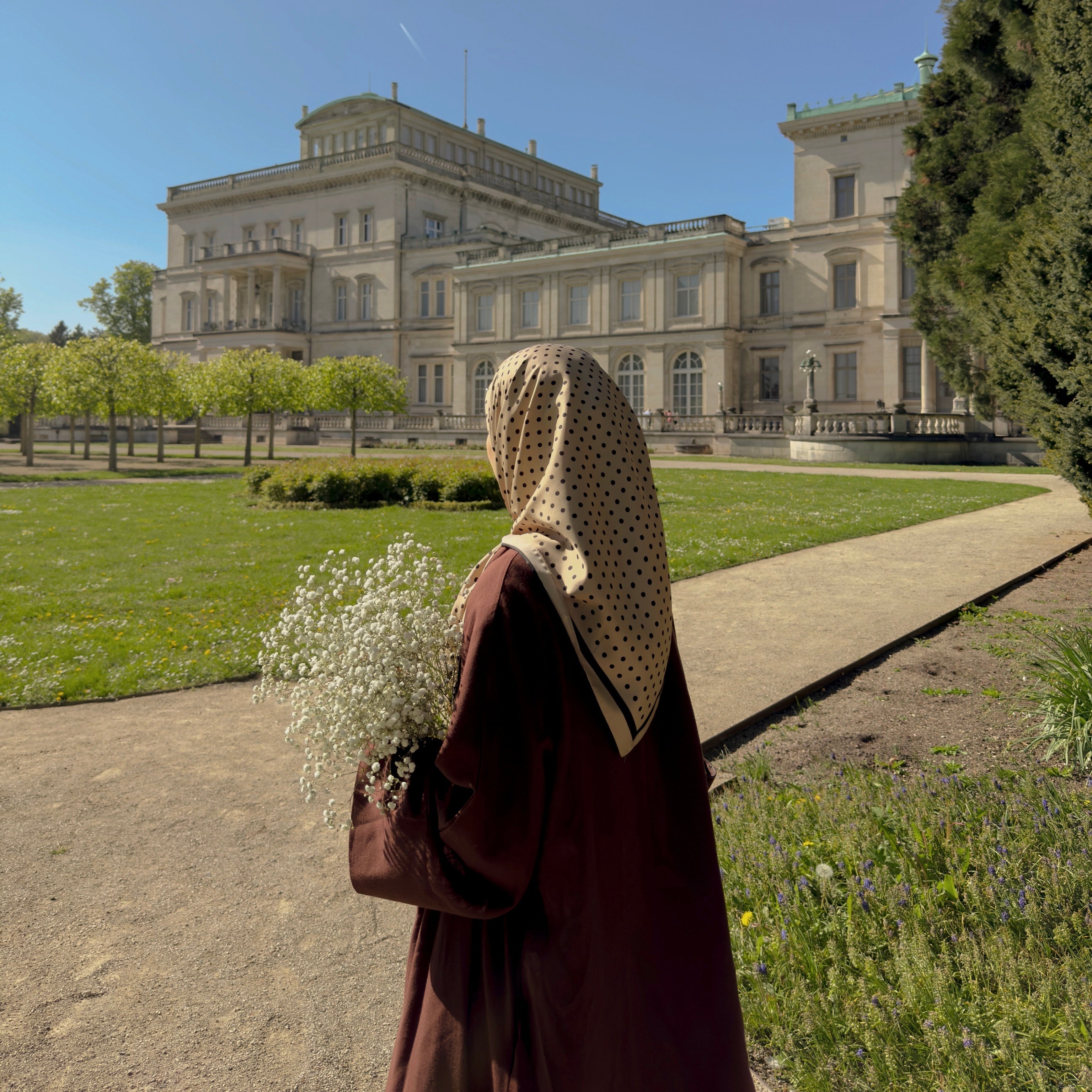 Person in a brown outfit with a headscarf standing in front of a large building with a green lawn and trees.
