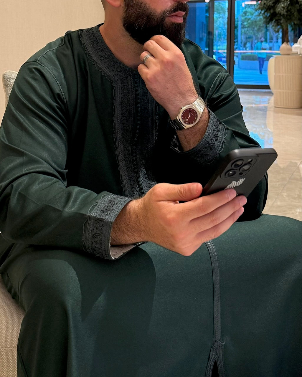 A man wearing a dark green traditional long-sleeve robe (thobe) and holding a black smartphone, sitting in a modern indoor setting.