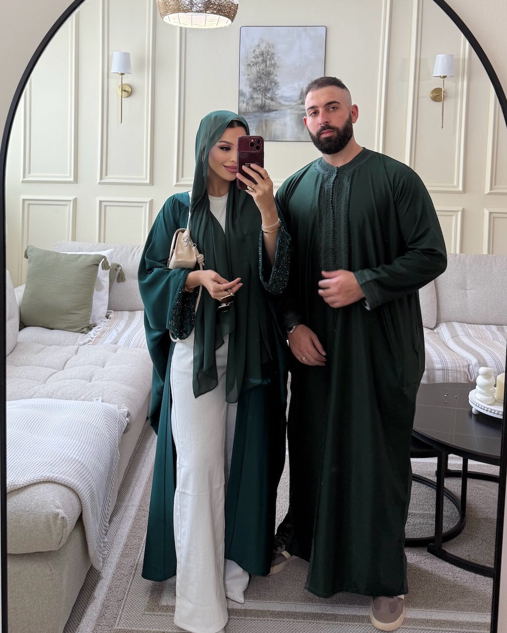 A couple taking a mirror selfie in a modern living room, wearing matching dark green traditional Arabic clothing. The woman wears a deep green abaya with a matching green hijab and white trousers, while the man wears a dark green thobe.