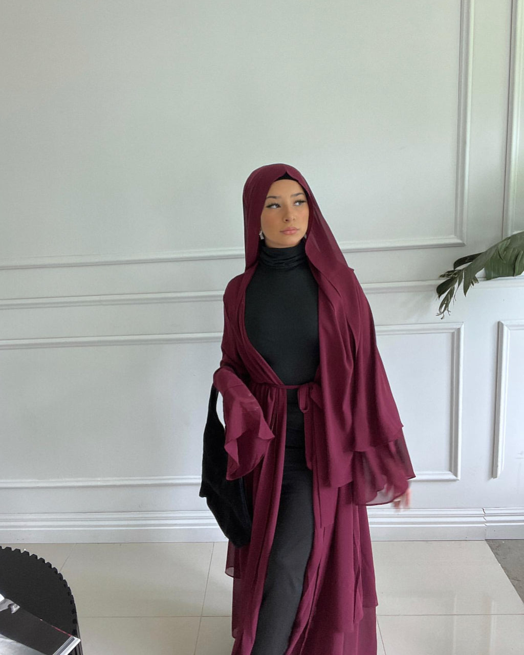 A woman wearing a long burgundy hijab and matching outer garment over a black turtleneck and dark pants, standing indoors against a white paneled wall.