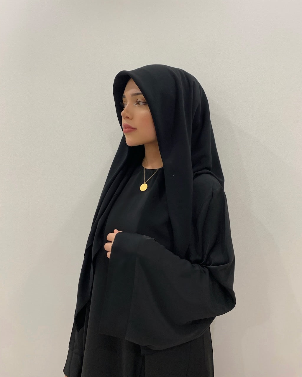 A woman wearing a black abaya and matching black hijab, styled with a delicate gold pendant necklace, standing against a plain light gray wall.