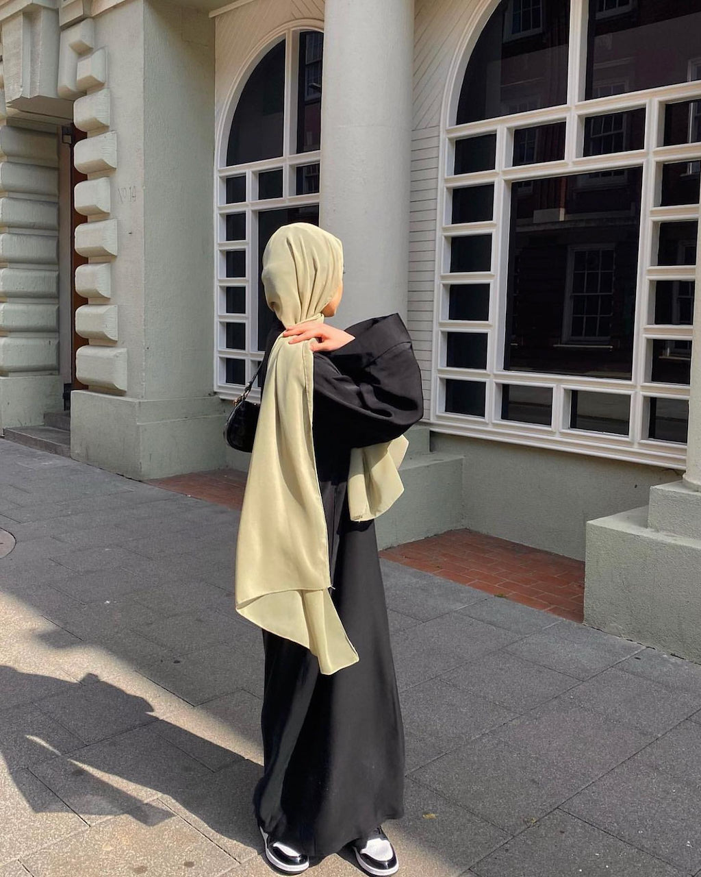 A woman wearing a beige hijab and a black abaya with a long black skirt stands on a sidewalk in front of a building with large white-framed windows.
