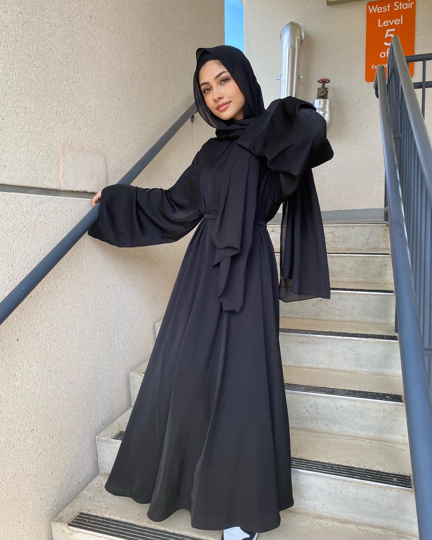 Woman in a black abaya standing on stairs with a neutral wall background