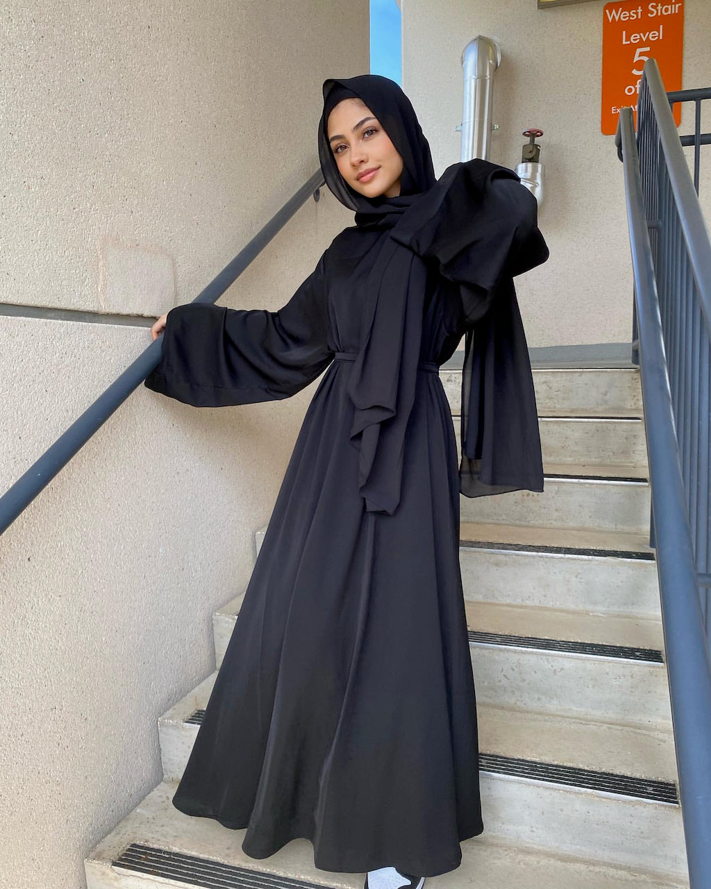 A woman wearing a long black abaya with a matching hijab stands on outdoor concrete stairs, holding a dark metal handrail. The abaya has a high ruffled collar and a self-tie waist.
