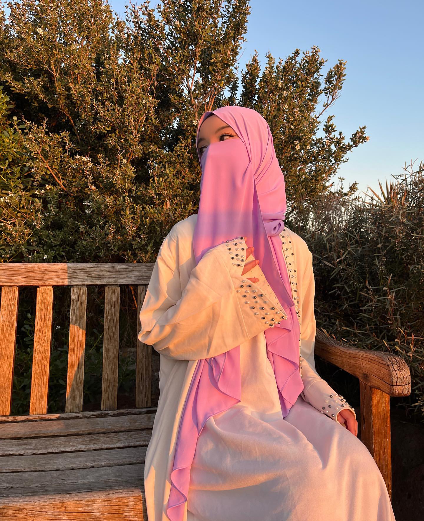 Person wearing a pink hijab and white outfit sitting on a wooden bench with trees in the background.